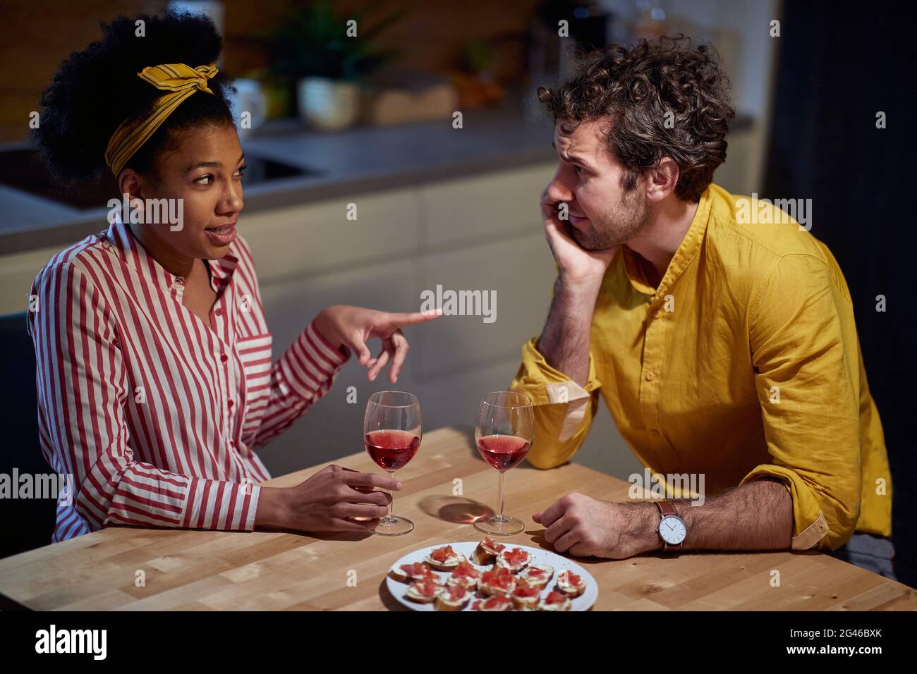 young adult afro-american female having a conversation with caucasian male, pointing with index finger at him, drinking wine, sitting at the table Stock Photo
