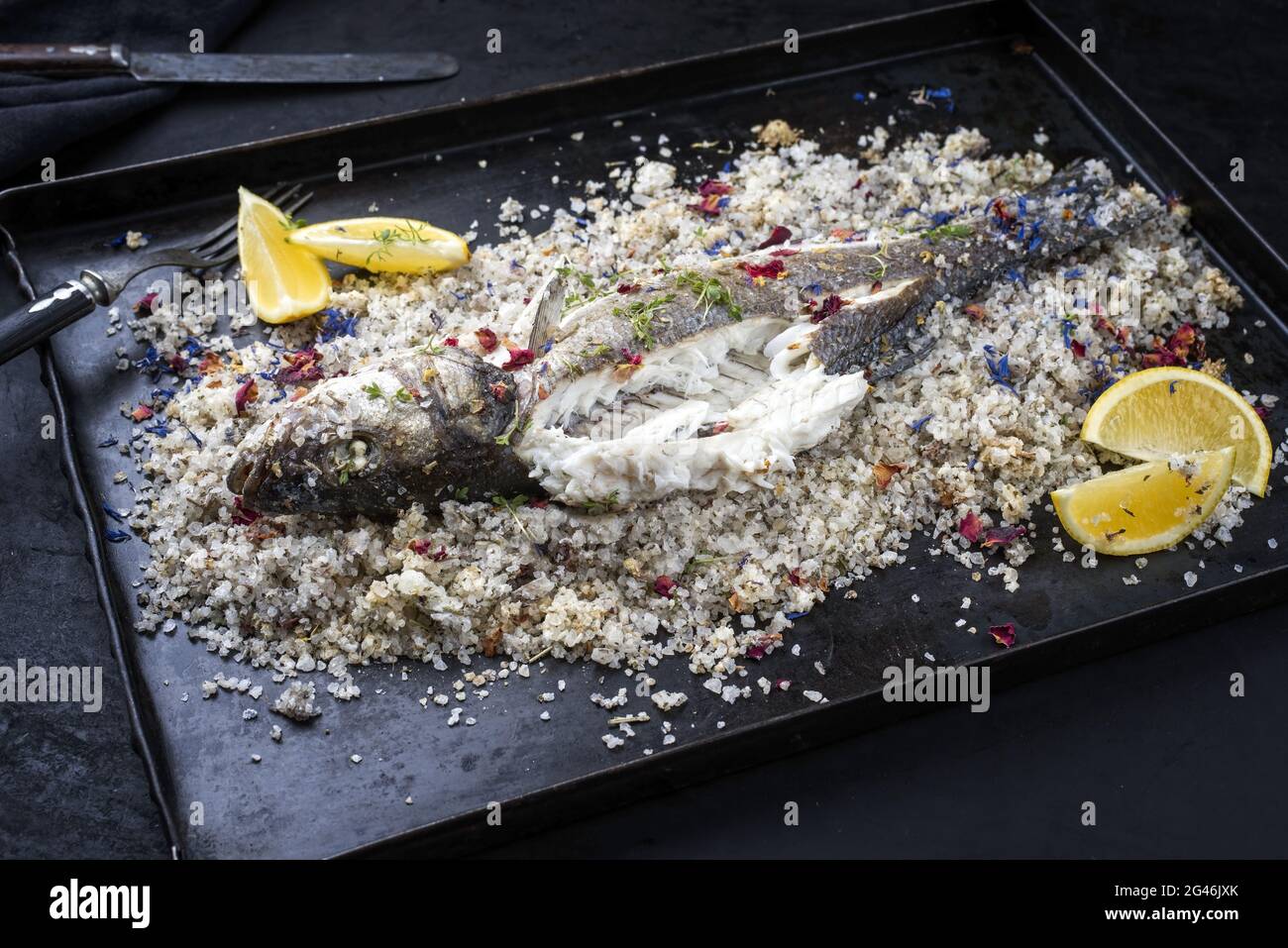 Traditional Atlantic Loup de Mer bedded into sea salt served with lemon slices and spices as close-up on a rustic metal tray Stock Photo
