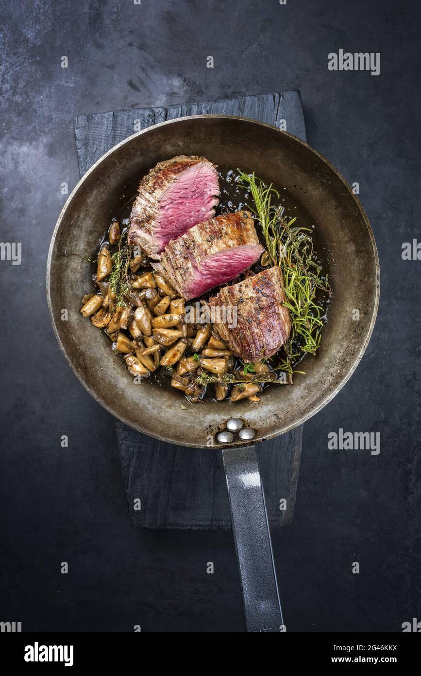 Fried dry aged beef fillet steak natural with king trumpet mushroom and herbs offered as top view in a rustic frying pan Stock Photo