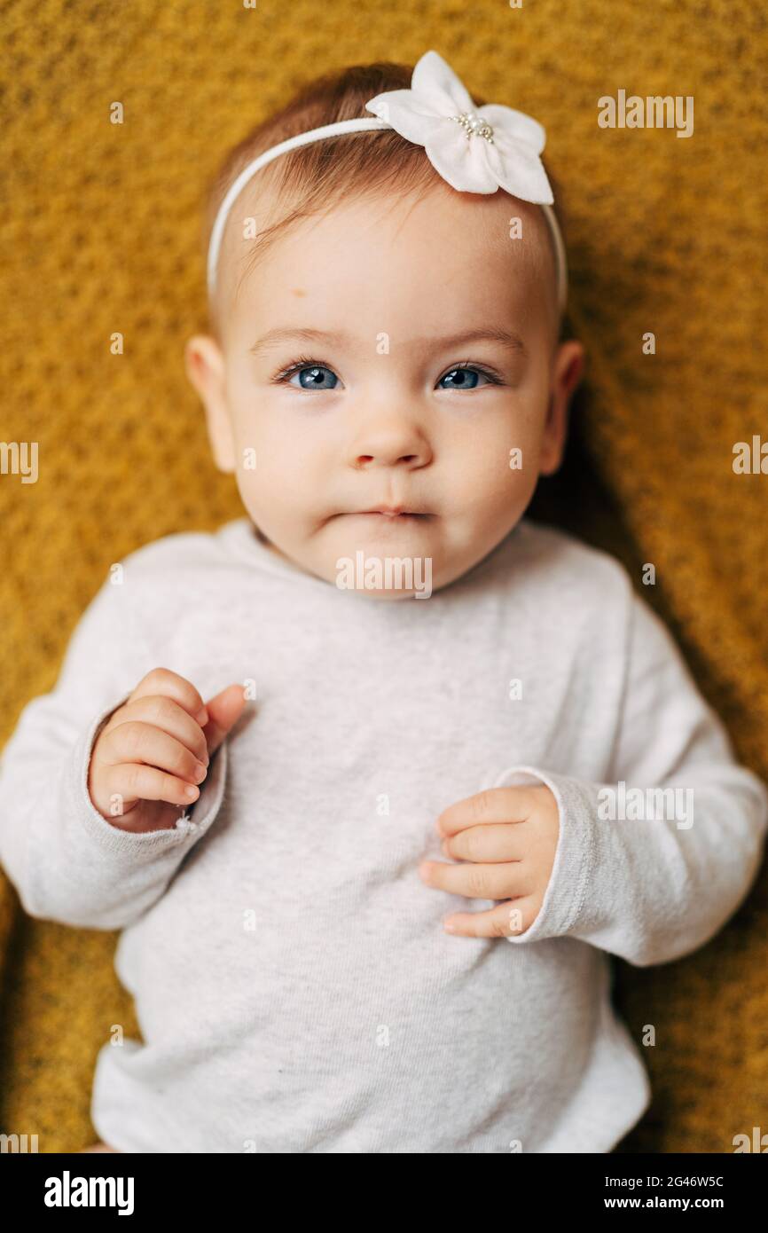 Pensive baby girl with a flower on her head in a white bodysuit lies on a yellow blanket, clenching her fists on her tummy Stock Photo