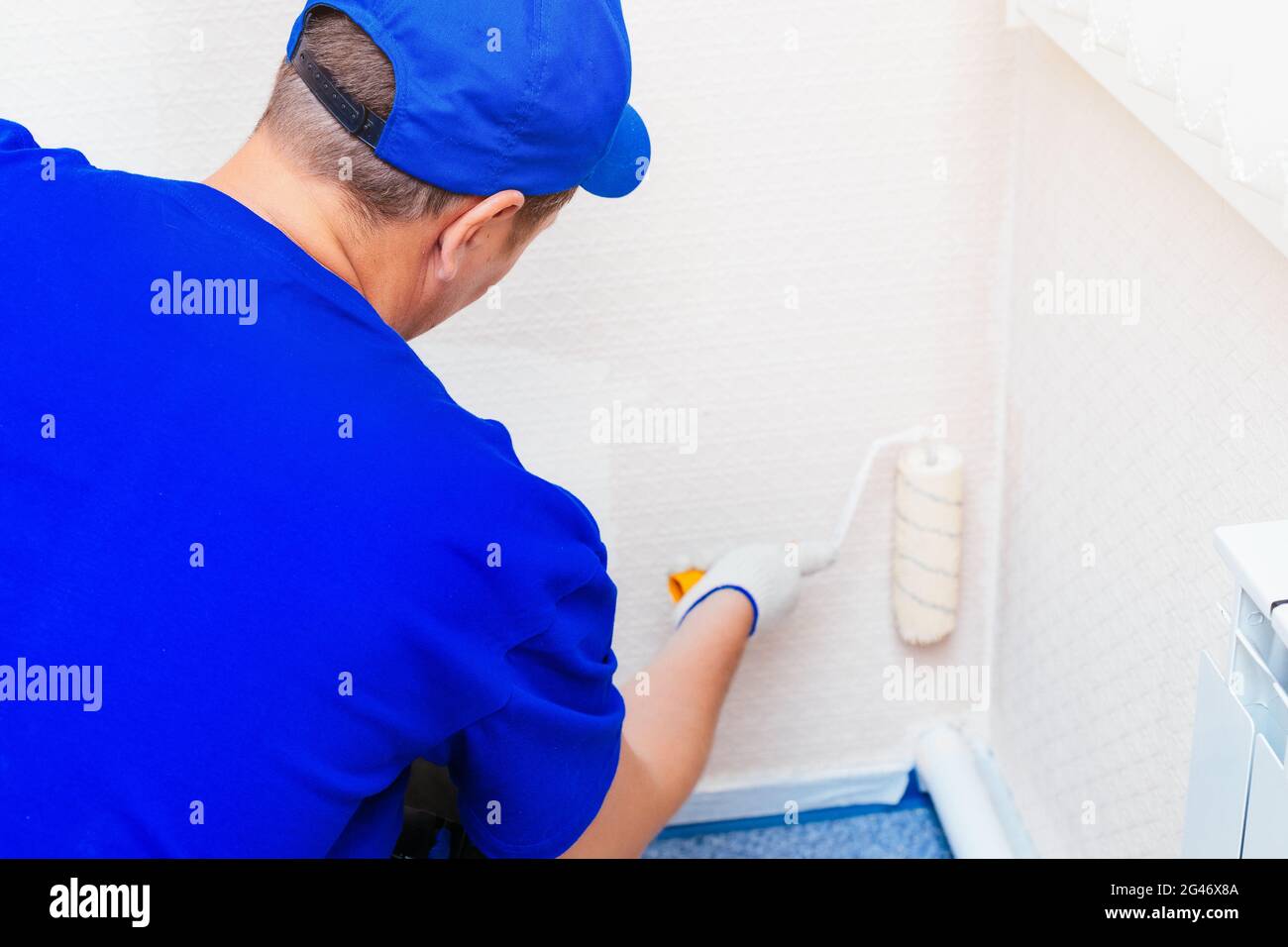 A painter in a cap and gloves paints the wall with a paint roller. Portrait of a young worker in uniform. Stock Photo