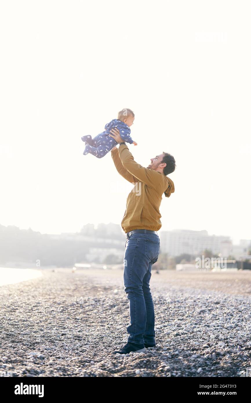 Smiling dad in a brown jacket and jeans throws a small child in a blue overalls high on his arms while standing on a pebble beac Stock Photo