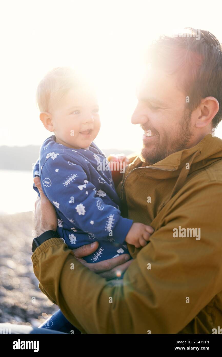 Smiling dad in a brown jacket sits and holds in his arms a small smiling child in a blue overalls. Sun shines brightly. Close-up Stock Photo