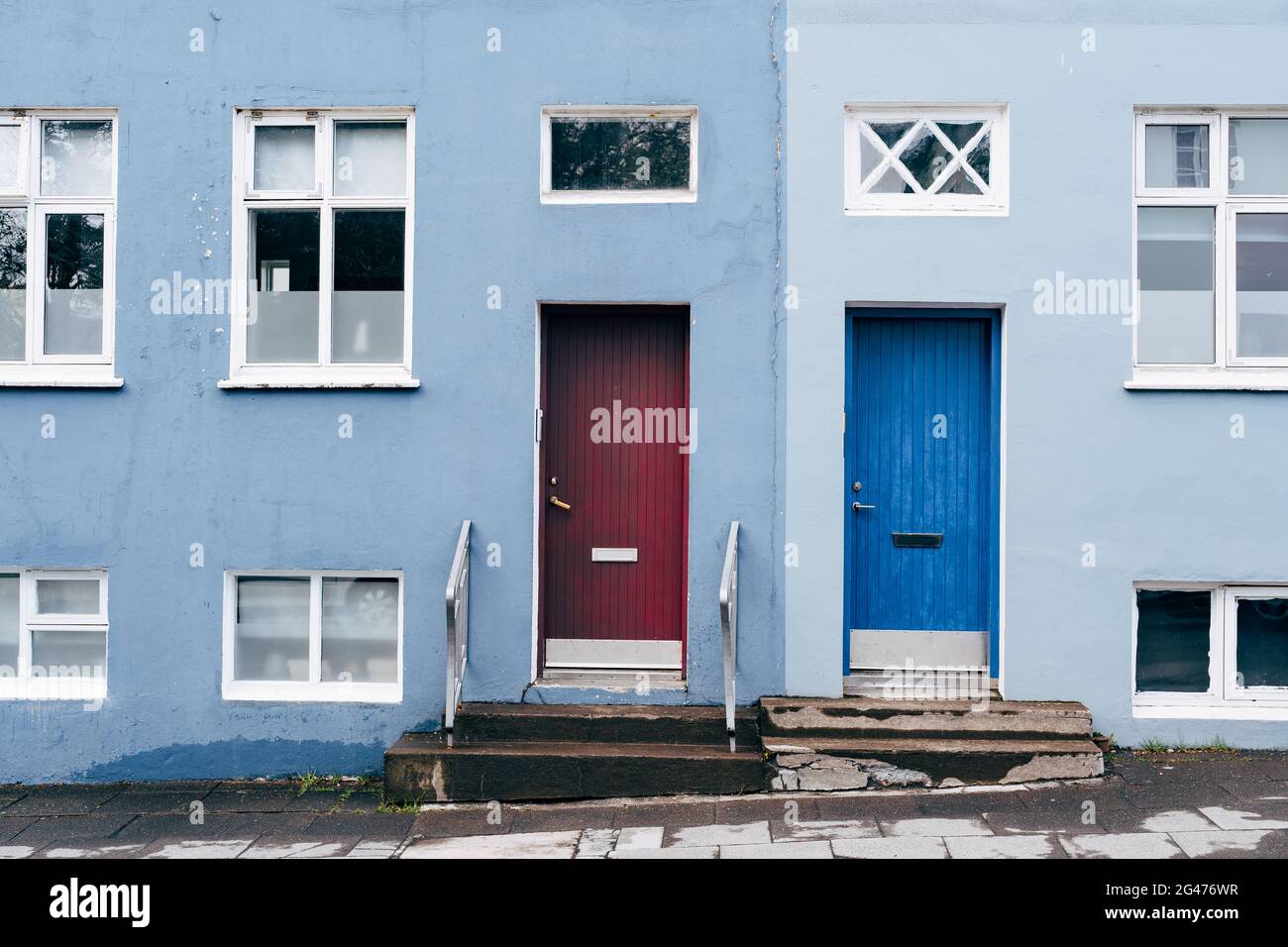 A two-tone building in blue and light blue with two rectangular doors in blue and burgundy and windows of different sizes. Stock Photo