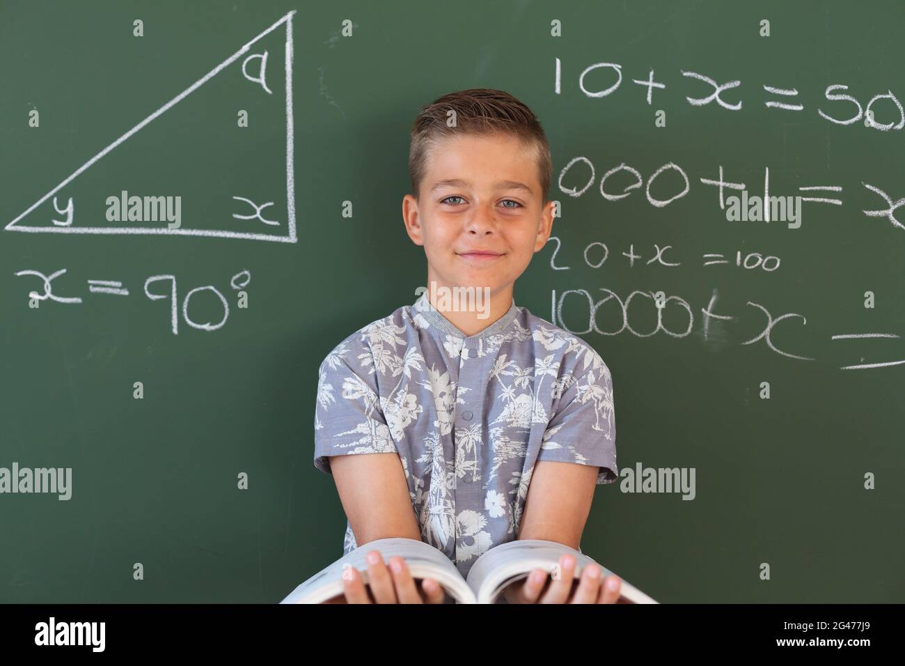 Portrait of happy caucasian boy standing at chalkboard in maths lesson classroom holding schoolbook Stock Photo