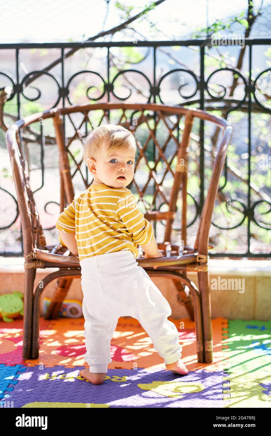 Kid stands on the balcony near the wicker chair, turning his head back Stock Photo