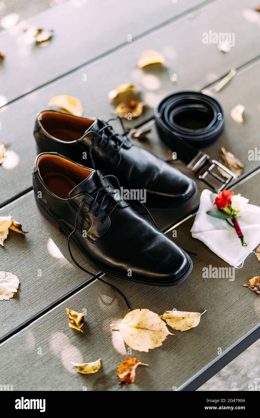 Black shoes of a man with untied laces on a wooden texture with a belt and groom's boutonniere on a white pocket square. Stock Photo