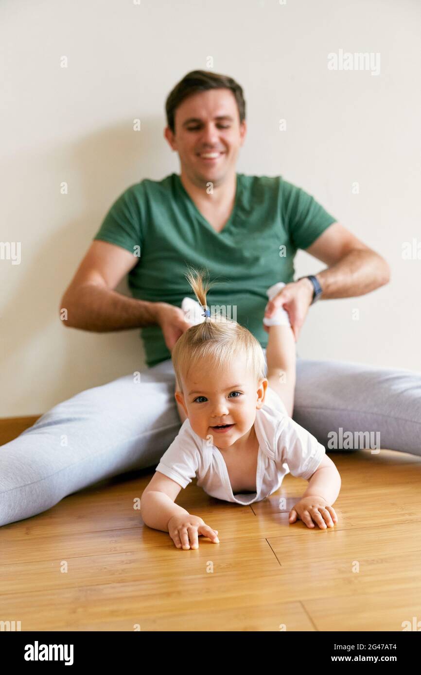 Smiling dad sits on the floor and pulls the crawling cute baby by the