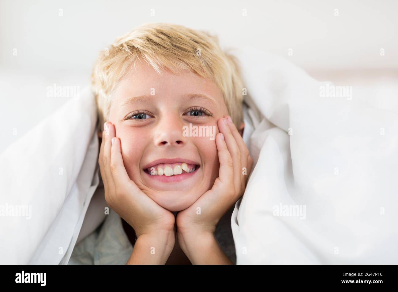Portrait of smiling boy lying under bed sheet in bedroom Stock Photo