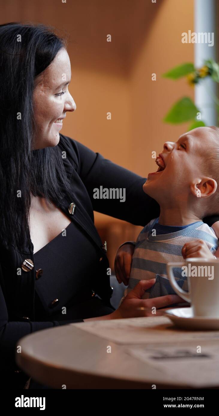 Close up portrait of a little boy with special needs and mom laughing at a table in a cafe, lifestyle. Mom's love for her child, inclusion.Happy disab Stock Photo