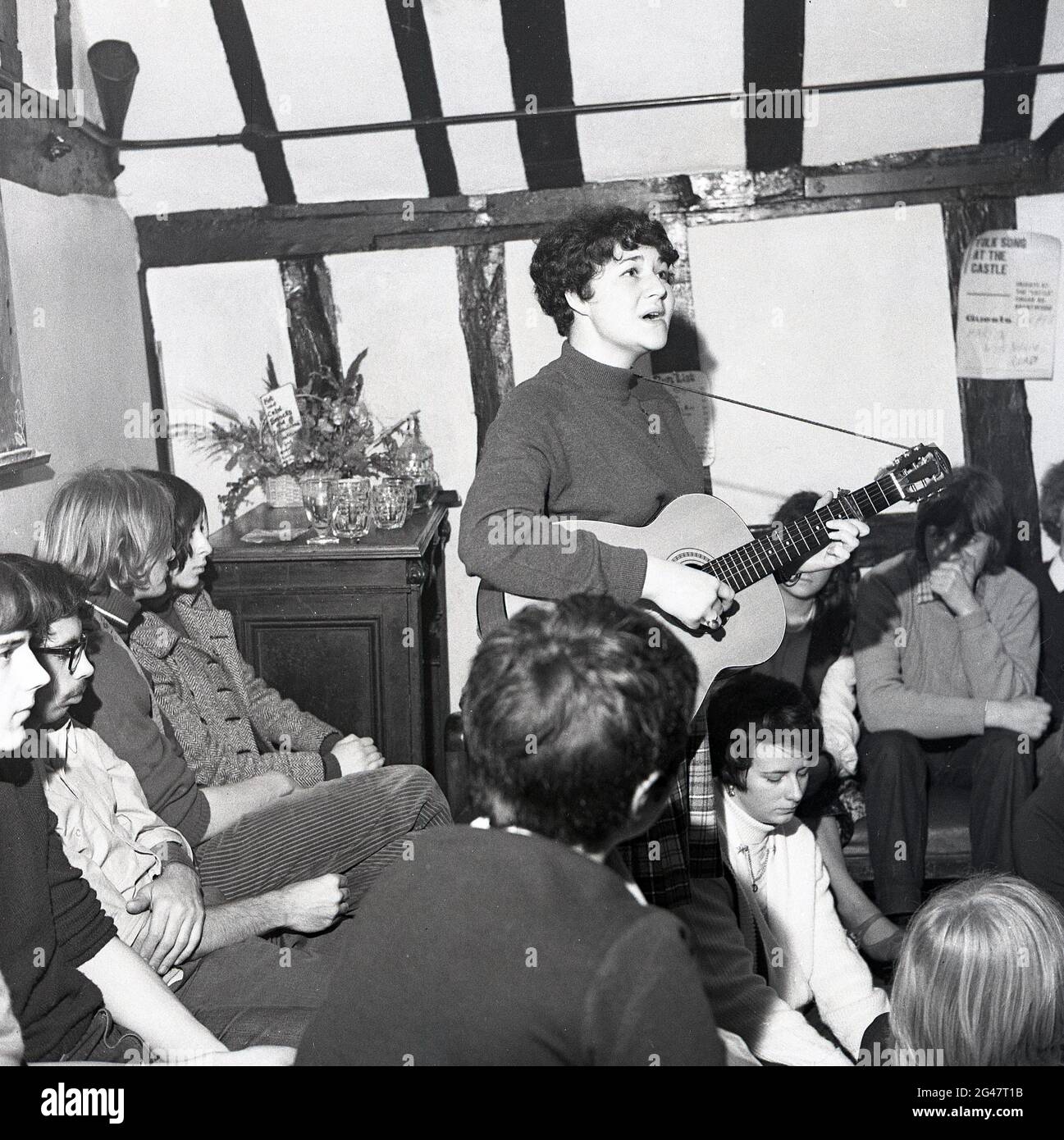 1970, historical, folk music, inside a room at a wooden beamed country pub, an audience listening to a young female folk singer playing a guitar and singing a song at the Castle Inn, Colchester, Essex, England, UK. Stock Photo
