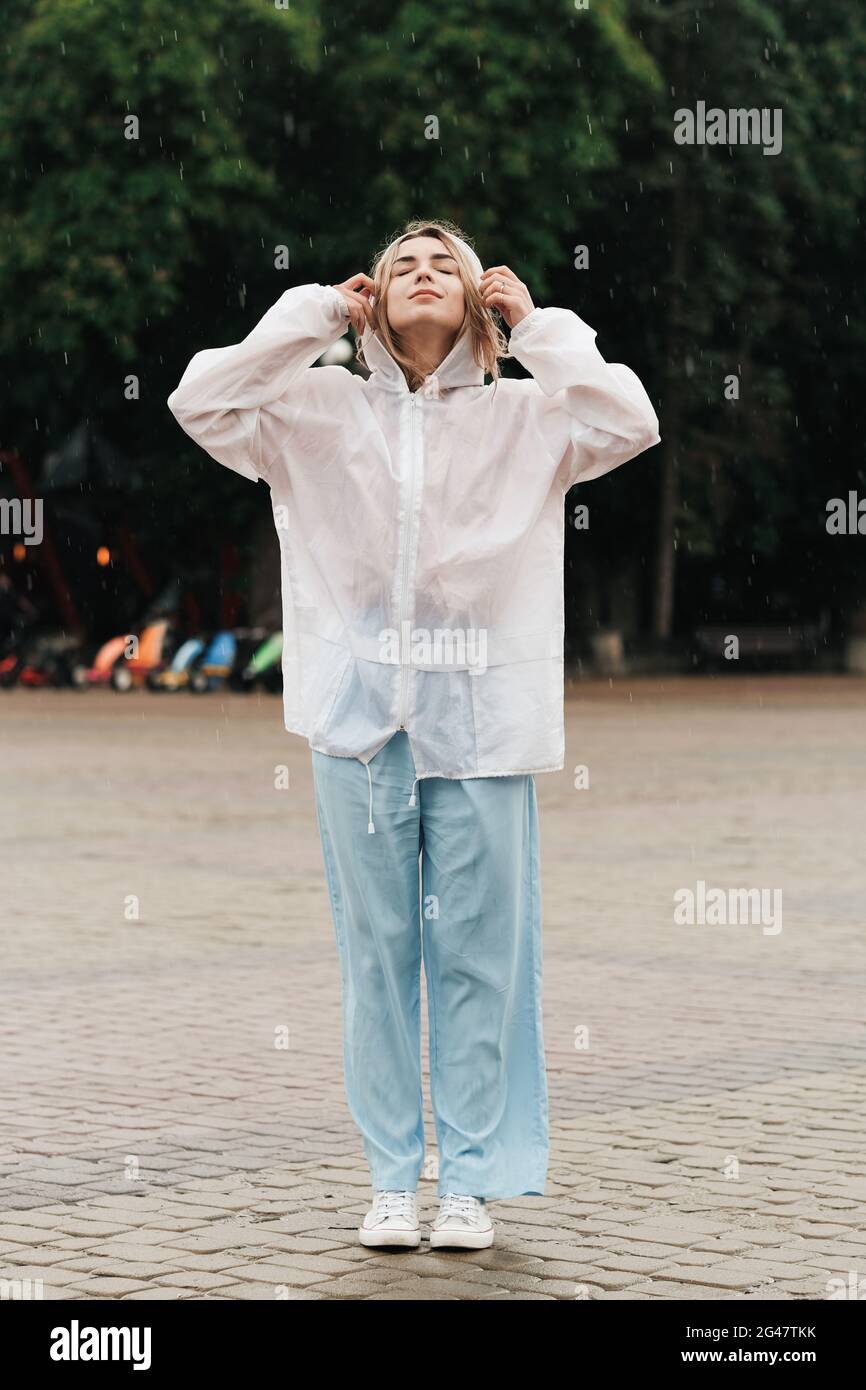 Portrait of Young Woman Wearing Raincoat While Standing on the Street at Rainy Day, Blonde Girl Dressed in Waterproof Cloak Having Fun Under Rain in Stock Photo