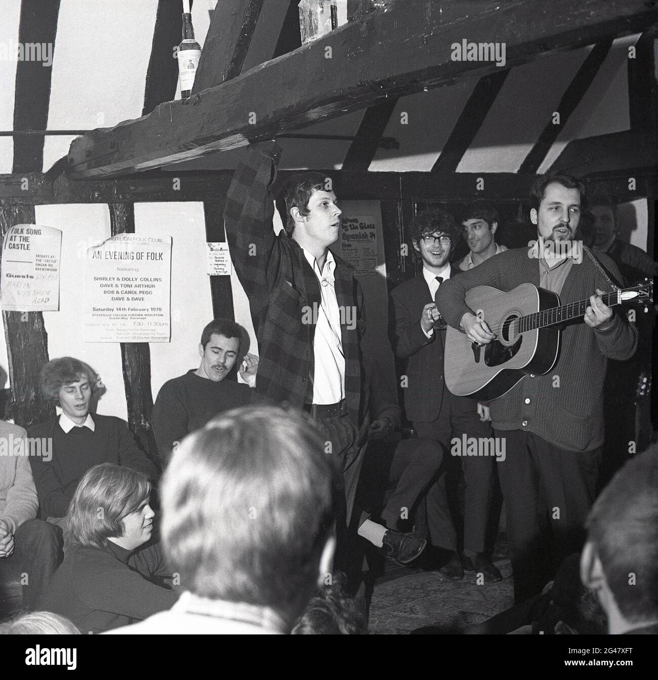 1970, historical, folk music, inside a wooden beamed room in a pub, an audience of young people listening to a folk musican, a man in a cardigan standing playing a guitar, The Castle Inn, Ongar Rd, Brentwood, Essex, England, UK. Publicity poster on the wall, giving notice of 'An Evening Of Folk' featuring sisters, Shirley & Dolly Collins, Dave & Toni Arthur, Bob & Carol Pegg, and Dave & Dave at the Stevenage College of Further Education, promoted by the Red Lion Folk Club. Stock Photo