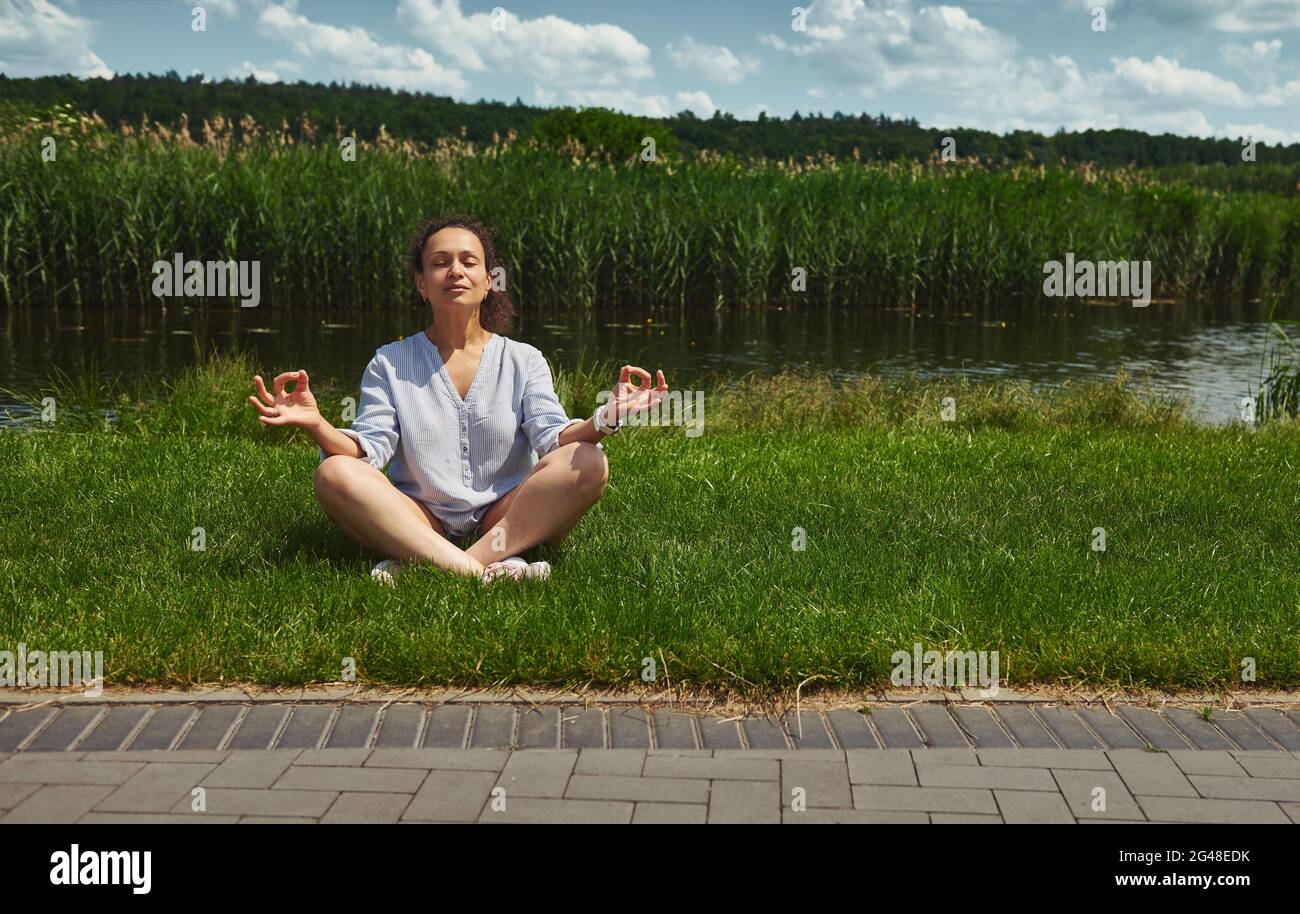 Young and serene woman sitting on the green grass in lotus position and ...