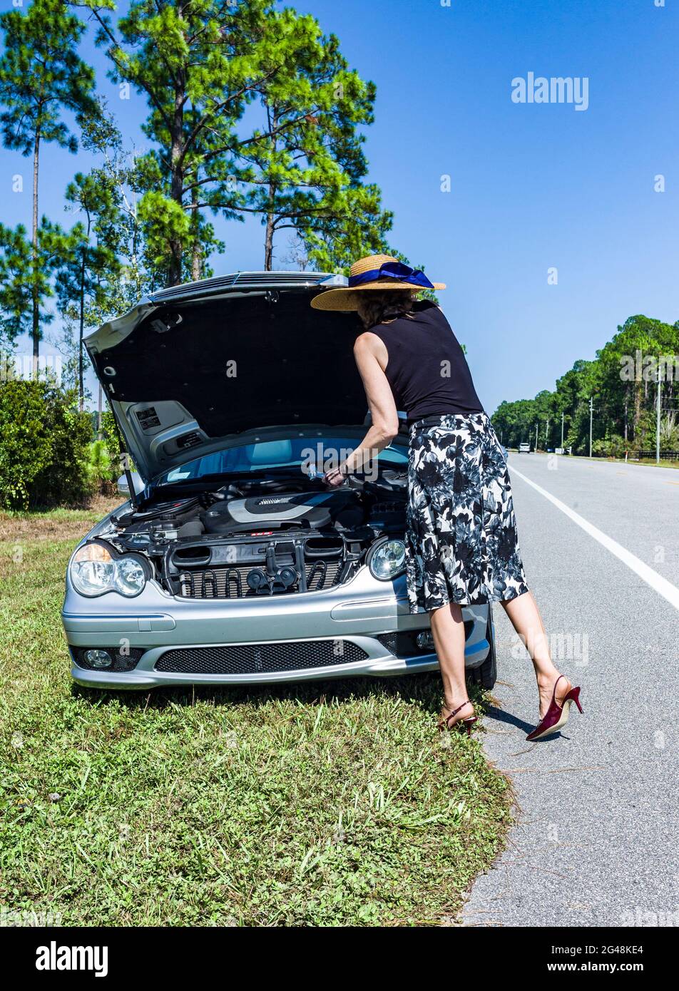 Well dressed anonomous woman holding a wrench while looking under the raised hood of car  parked on the side of the road. Stock Photo