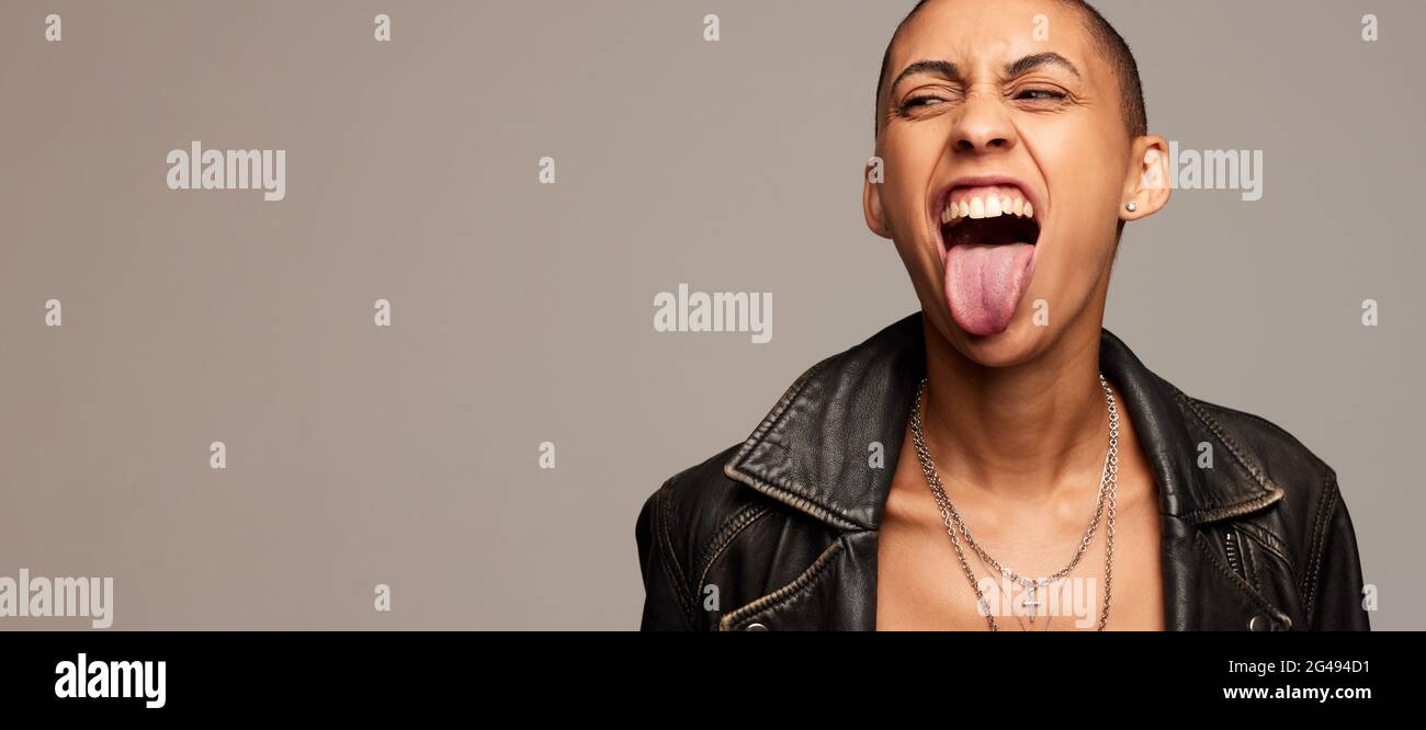 Excited androgynous woman sticking out her tongue on gray background. Expressive woman with shaved head looking away at copy space. Stock Photo