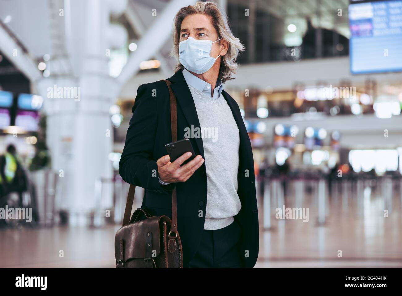 Man wearing facemask with handbag and mobile phone on airport. Male business traveler in transit area at an international airport. Stock Photo