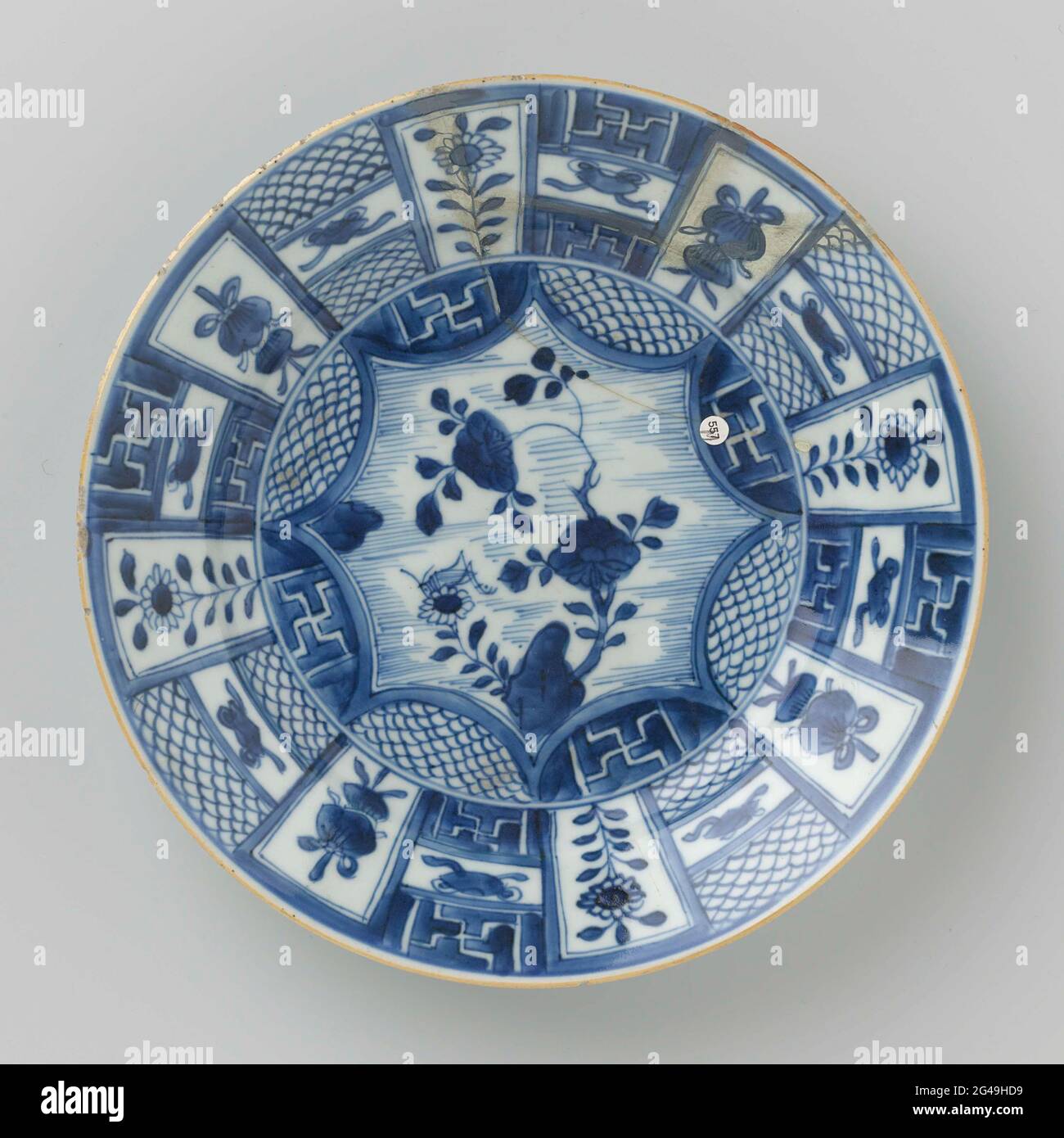 Plate With Panel Decoration With Flower Sprays and Precious Objects. Porcelain plate with raised wall and sloping edge, painted in underglaze blue. On the flat a pointed medallion with flower branches and insect at a rock, half circles around the medallion filled with alternating a scaled motif and Swastika in reserve; the edge with a trade decoration with alternating a flower branch or lucky symbol (parasol) and a three-piece compartment with valuables (pearl, rhombus) above and-under or a schub pattern or a swastika in reserve; The back with a continuous flower branch. Plate has been broken. Stock Photo