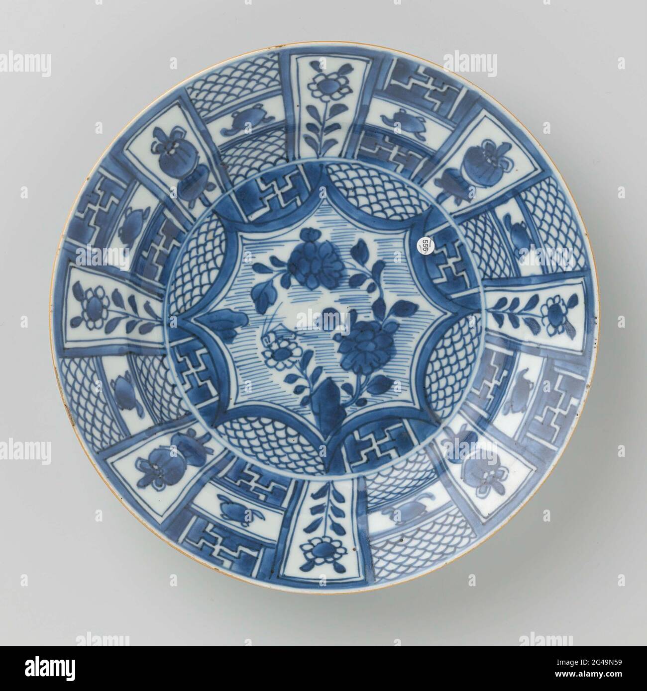 Plate With Panel Decoration With Flower Sprays and Precious Objects. Porcelain plate with raised wall and sloping edge, painted in underglaze blue. On the flat a pointed medallion with flower branches and insect at a rock, half circles around the medallion filled with alternating a scaled motif and Swastika in reserve; the edge with a trade decoration with alternating a flower branch or lucky symbol (parasol) and a three-piece compartment with a pearl above and-under or a schub pattern or a swastika in reserve; The back with a continuous flower branch. Blue White. Stock Photo