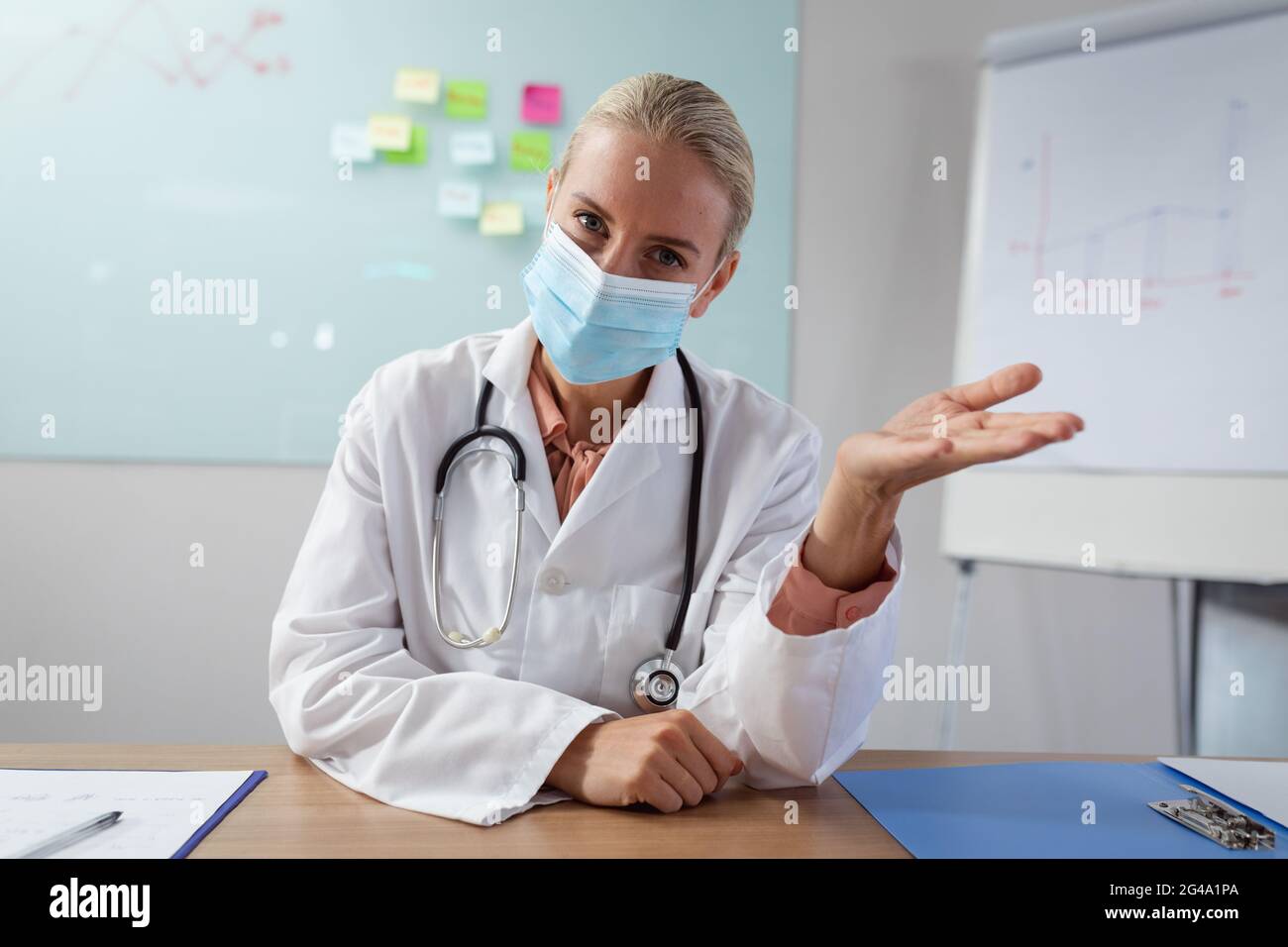 Caucasian female doctor wearing face mask sitting at desk in office gesturing during video call Stock Photo