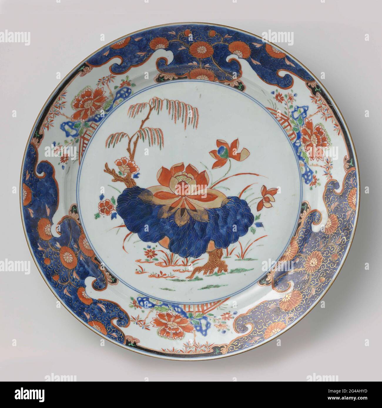 Dish with Large Lotus, Weeping Willow and Plants. Dish of porcelain, painted in underglaze blue and on the glaze blue, red, green, black and gold. On the flat a large lotus plant and a weeping willow; On the wall and edge four groups with a rock, fence and flowering plants such as Japanese aster, peony and bamboo; above these groups each time a band with lotus drinks against a black soil; Between the groups a box with curl work and chrythanity against a blue ground. Chinese Imari. Stock Photo