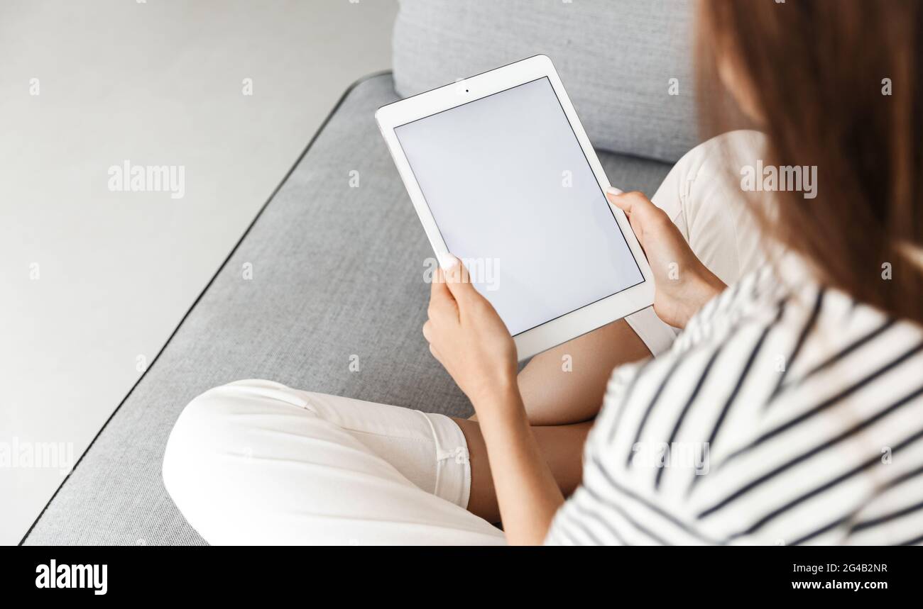 Cropped shot of woman, hands holding digital tablet with white screen, mockup promotion empty space on display, sitting with crossed legs on sofa at Stock Photo