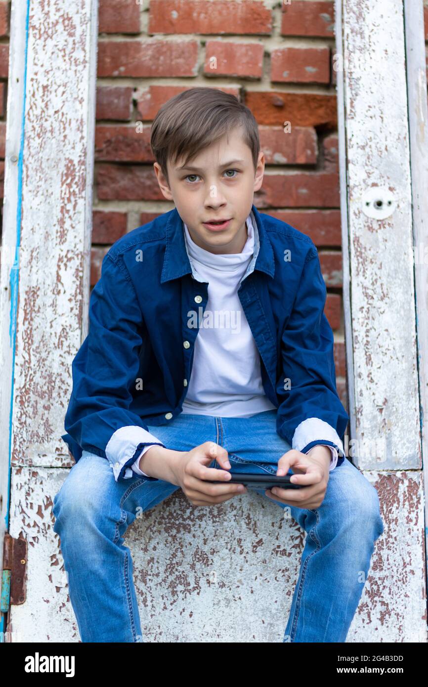 A teenager boy in a blue shirt and jeans with a smartphone in his hands  sits on an old door against a brick wall background. Selective focus Stock  Photo - Alamy
