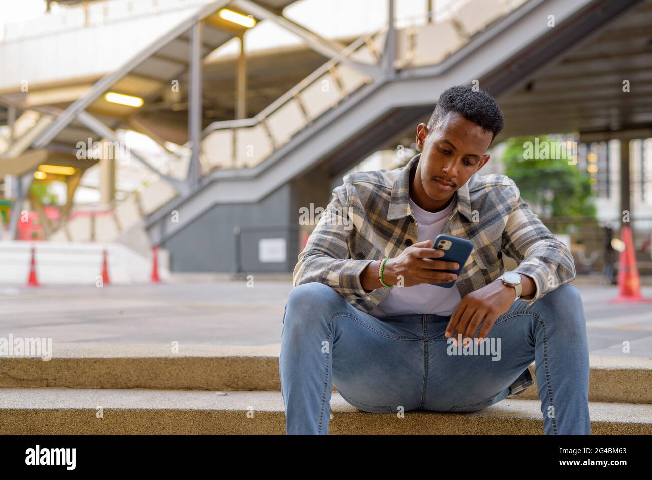Portrait of African black man sitting outdoors in city during summer using mobile phone horizontal shot Stock Photo
