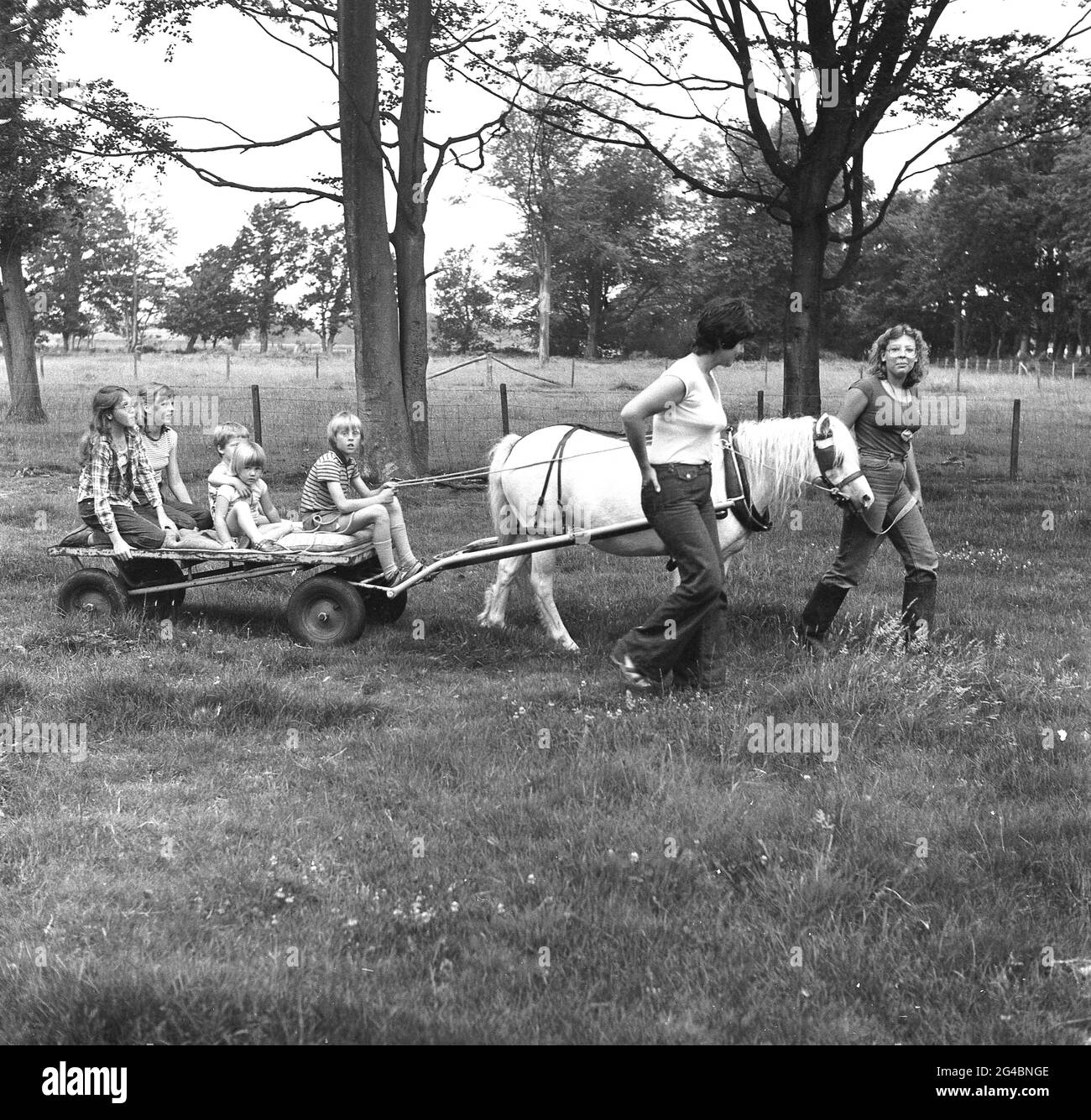 1980s, historical, holiday family fun on a farm, some young children having a ride on a small four-wheeled flat trolley or platform, pulled by a horse around a field, led by a female farm worker and a mother beside, Hordle, Hampshire, England, UK. Stock Photo