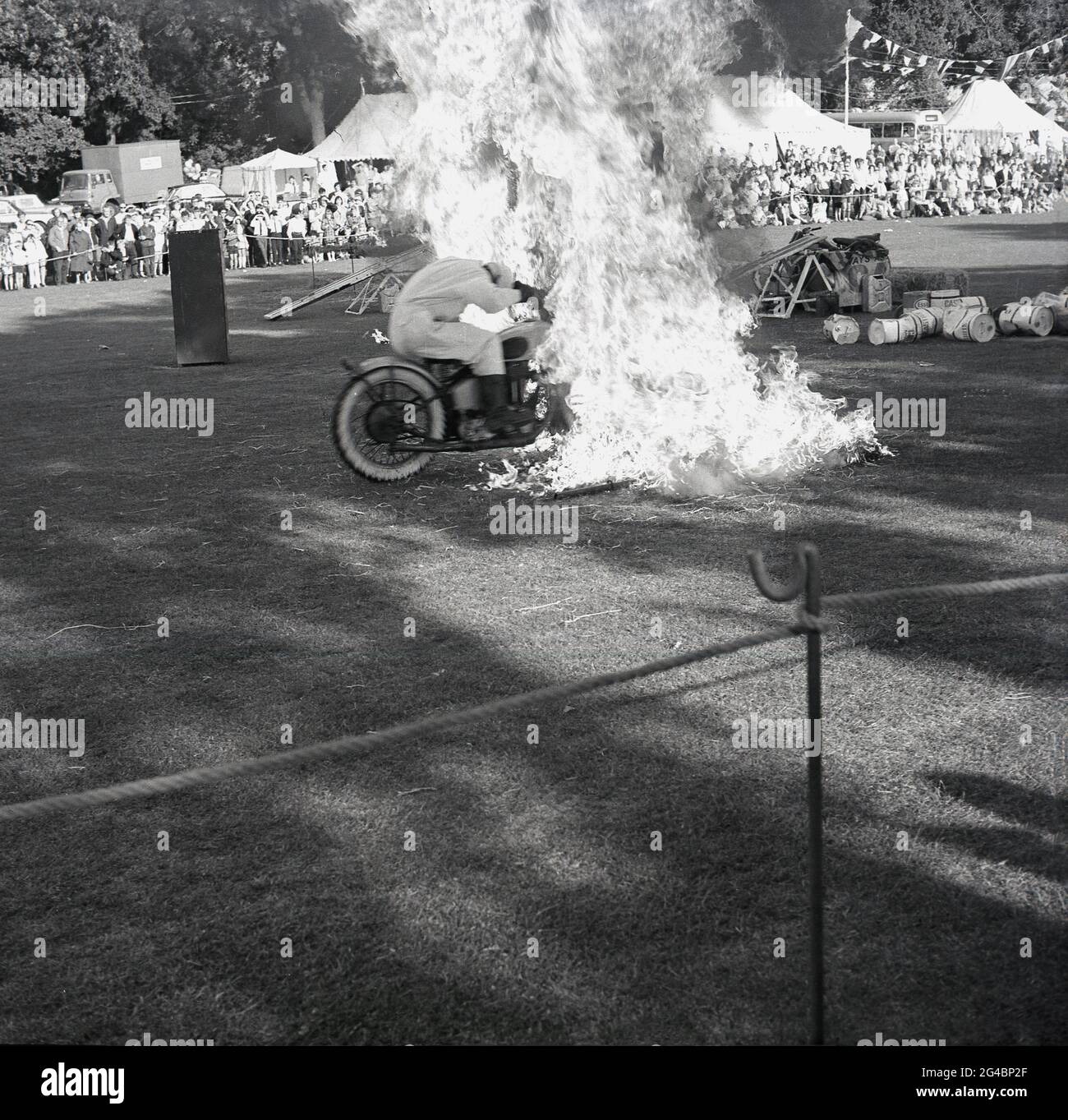 1960s, historical, outside in a field, at a county show or fete, spectators watching a motorcycle display, with one of the display team riding though fire, a high-risk, dramatic daredevil stunt, England, UK. Motorcycle acrobatic displays at outside carnivals and special events were popular in this era, with the Royal Signals, the 'White Helmets', an army display team, the most well-known. They had performed the first recorded stunt display in 1928 as the despatch riders who were a key part of the army's communications, needed to be highly skilled motorcyclists. Stock Photo