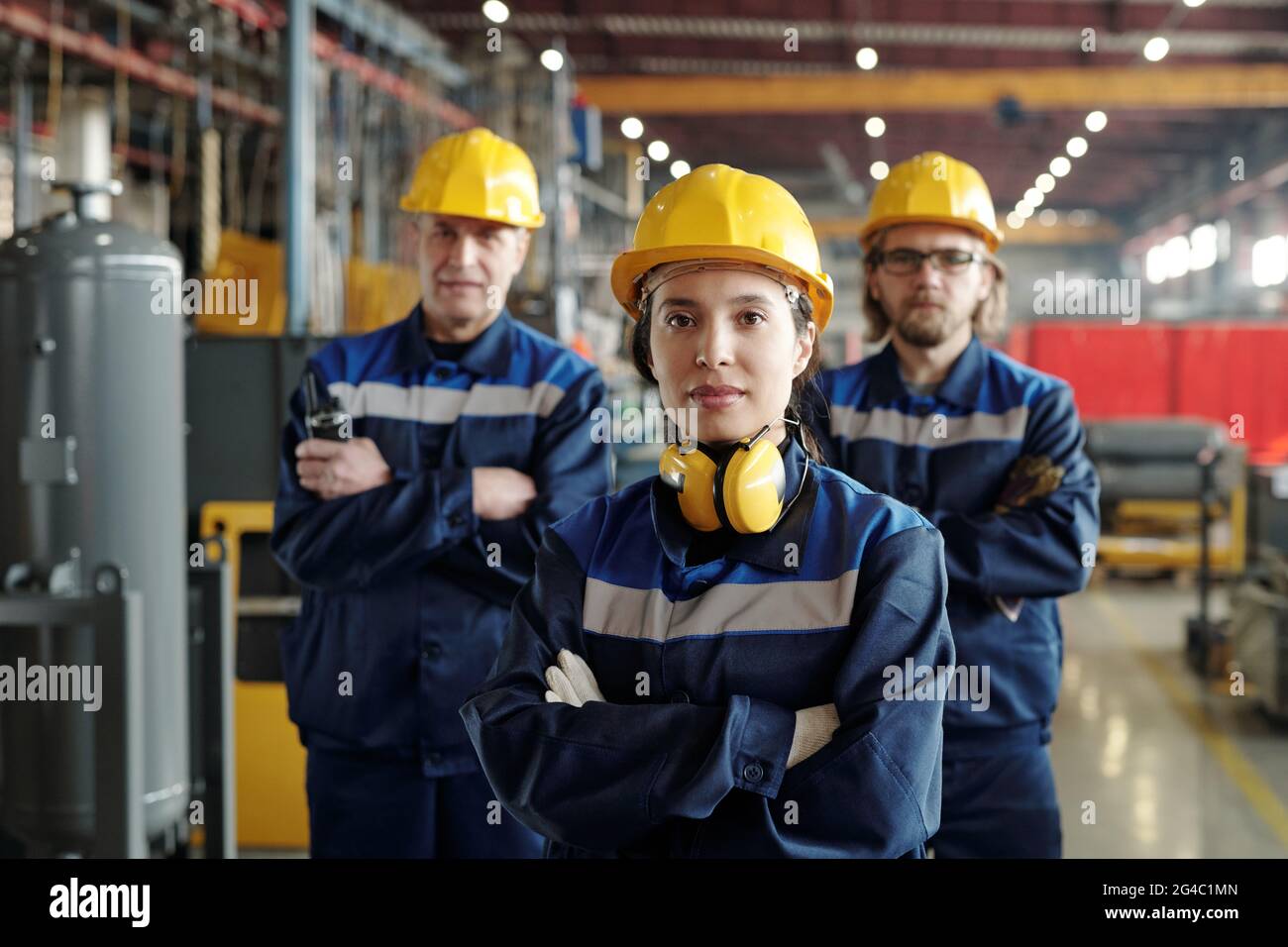 Portrait of content young female plant worker with sound-proof headphones standing with crossed arms against colleagues in compressor factory shop Stock Photo