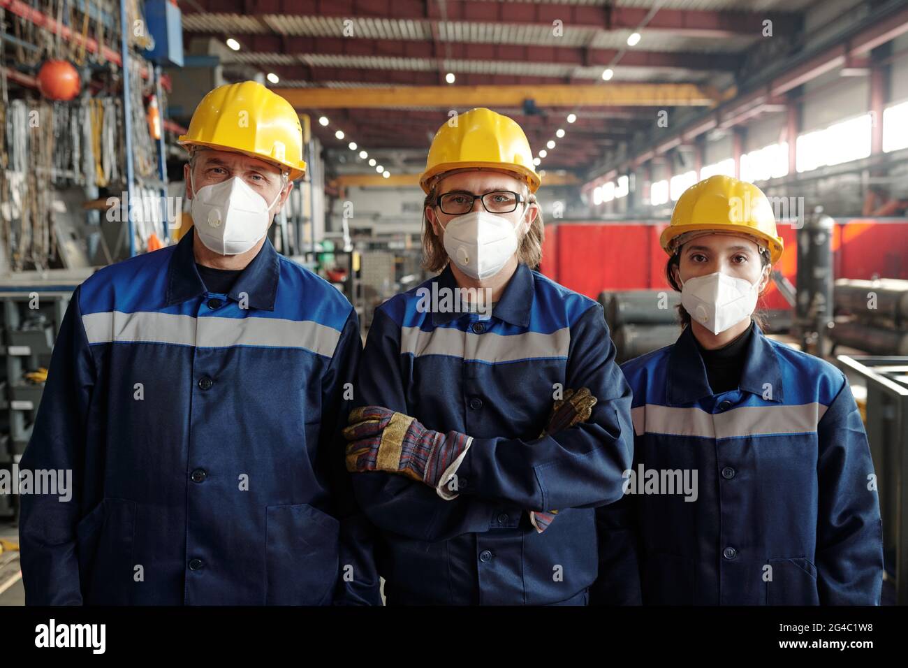 Portrait of serious confident manufacturing workers in hardhats and masks standing in line at modern factory Stock Photo