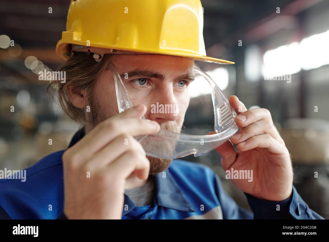 Close-up of confident young bearded factory worker in yellow hardhat putting safety goggles on face while preparing for work Stock Photo