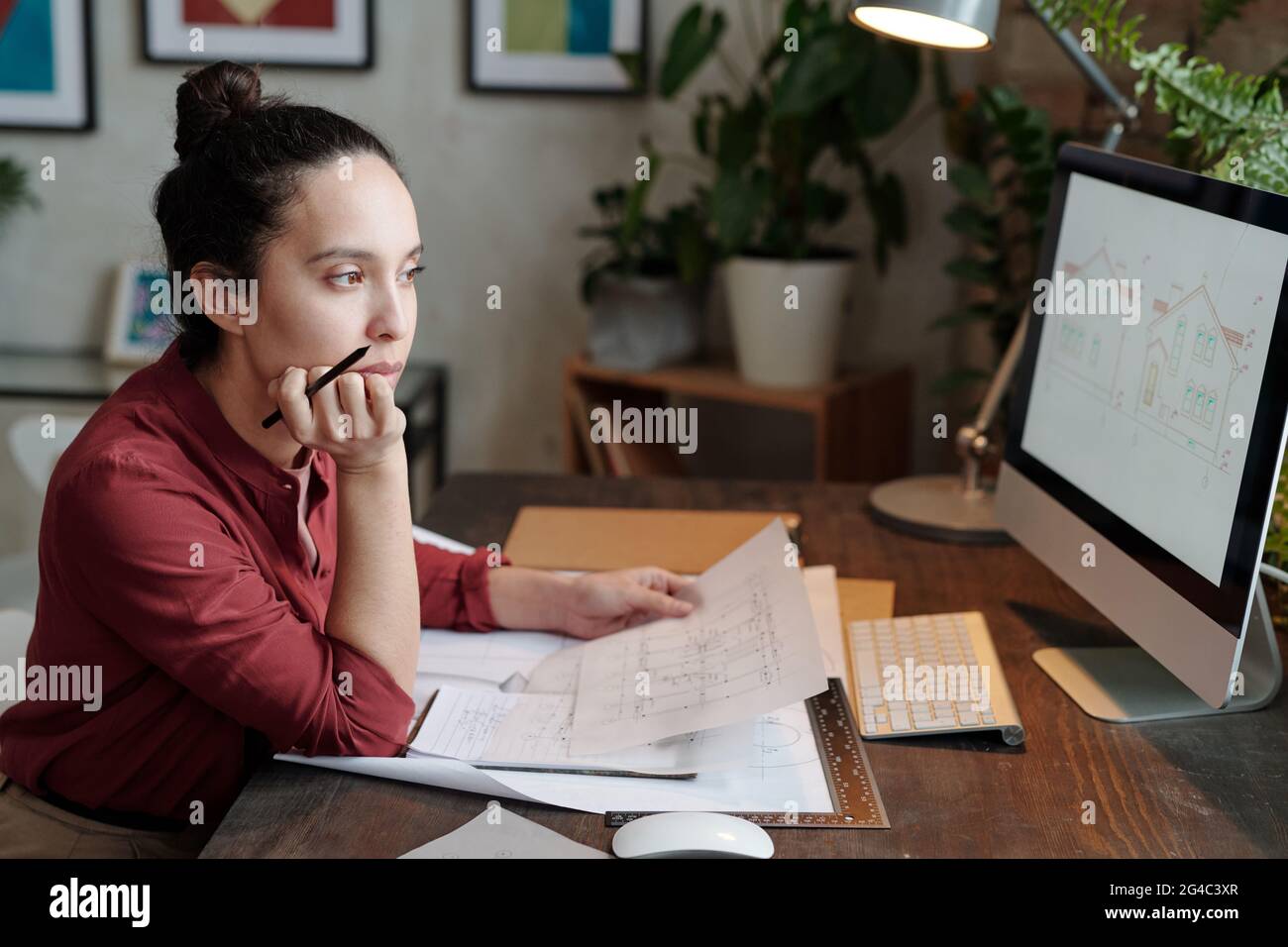Serious young mixed race woman with hair bun sitting at desk and holding house plan while analyzing design sketch on computer Stock Photo