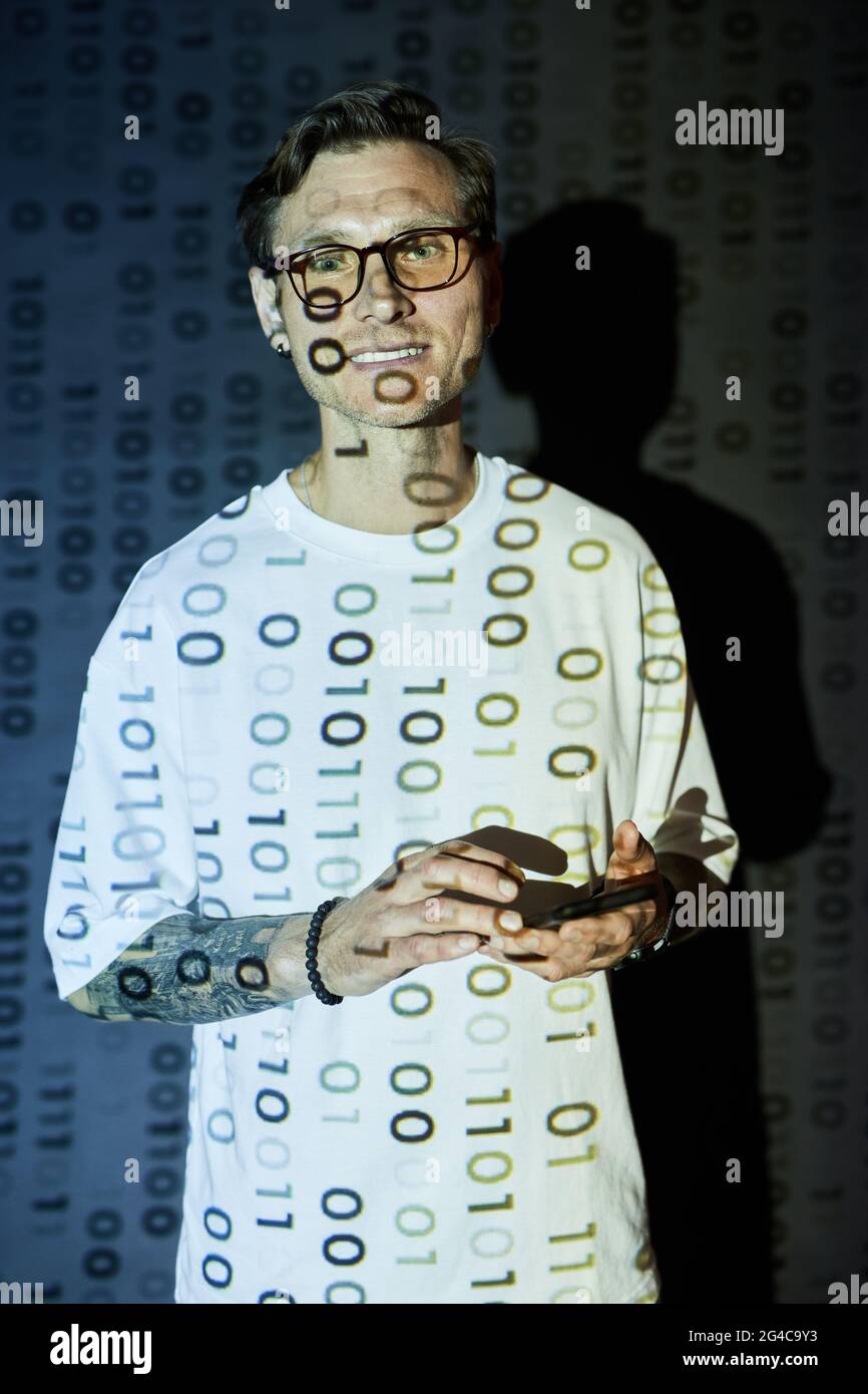 Portrait of young man in eyeglasses smiling at camera while using mobile phone in cyber security center Stock Photo