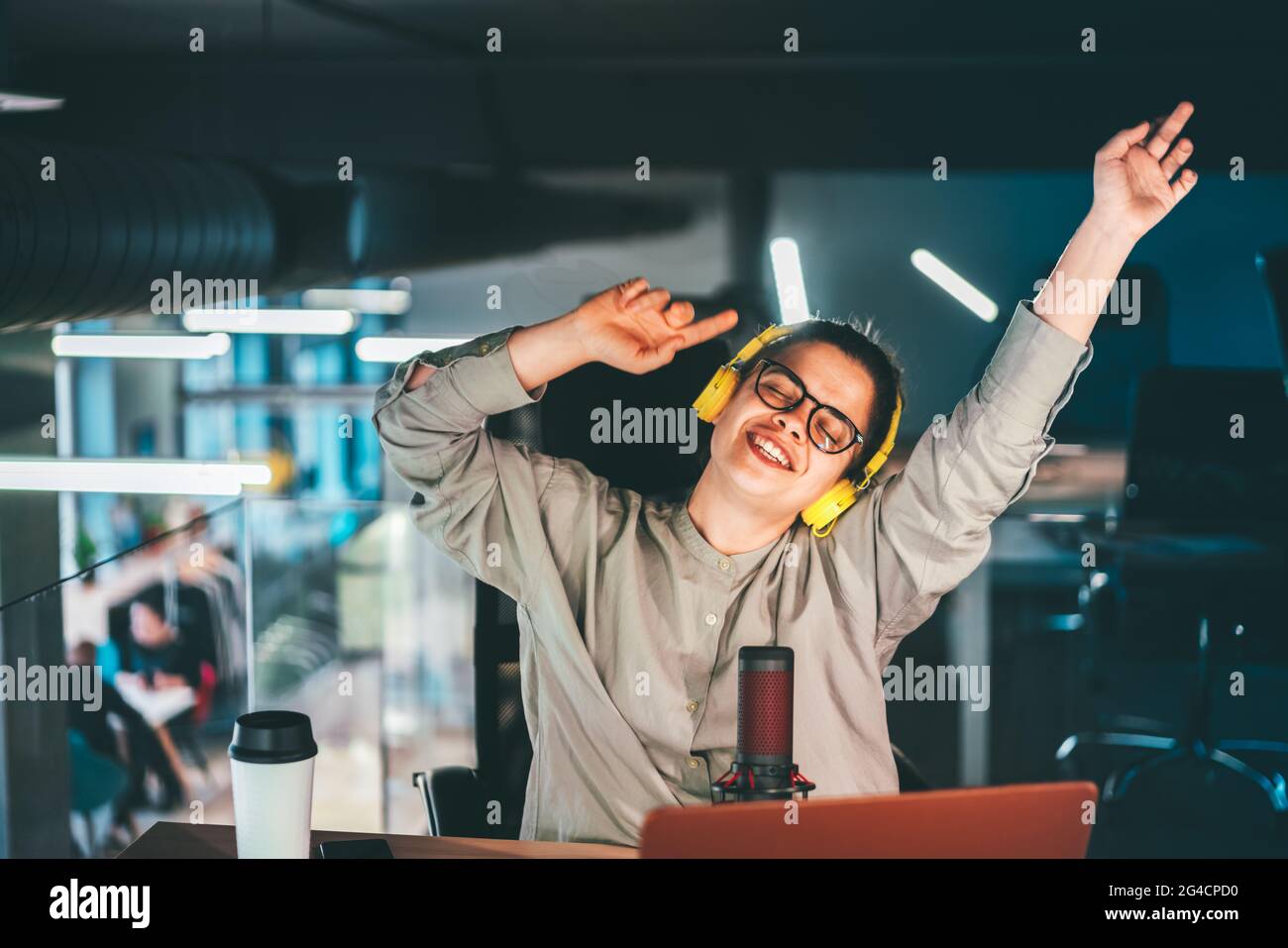Attractive young woman in her 30s wearing glasses and yellow headphones. Happy singing and dancing at her workplace with her hands raised up, portrait Stock Photo