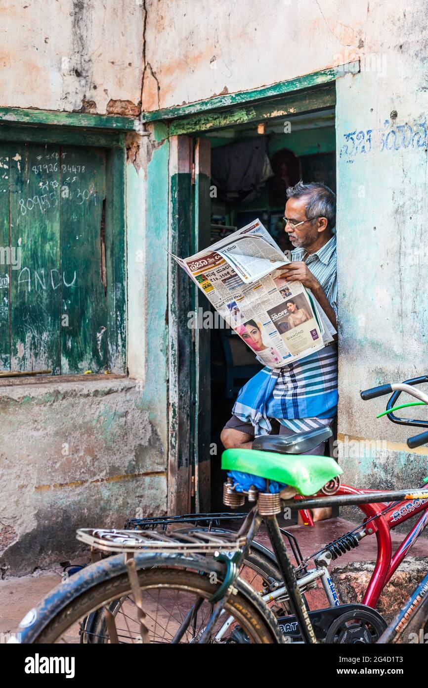 Indian gentleman with gray hair, spectacles and lunghi stands in doorway reading newspaper, Gokarna, Karnataka, India Stock Photo