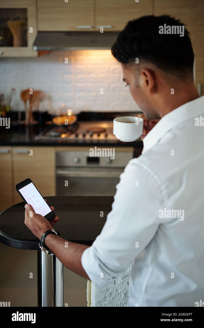 Young investor drinking cup of morning coffee in kitchen and checking price of bitcoin and predictions via appliction on smartphone Stock Photo