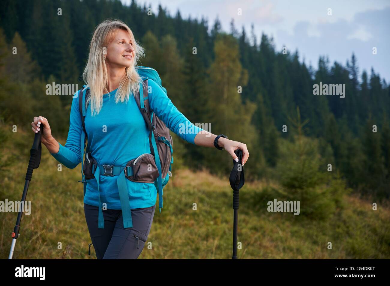 Joyful female traveler looking at beautiful mountains and smiling while holding trekking poles. Charming young woman hiker carrying backpack and holding hiking sticks while standing on grassy hill. Stock Photo