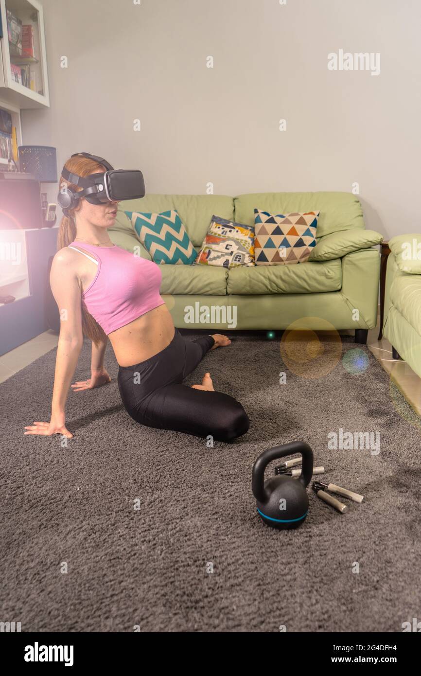 Attractive young red hair sportswoman training in living room of her beautiful house doing stretching with vr futuristic goggles - Ginger model Stock Photo