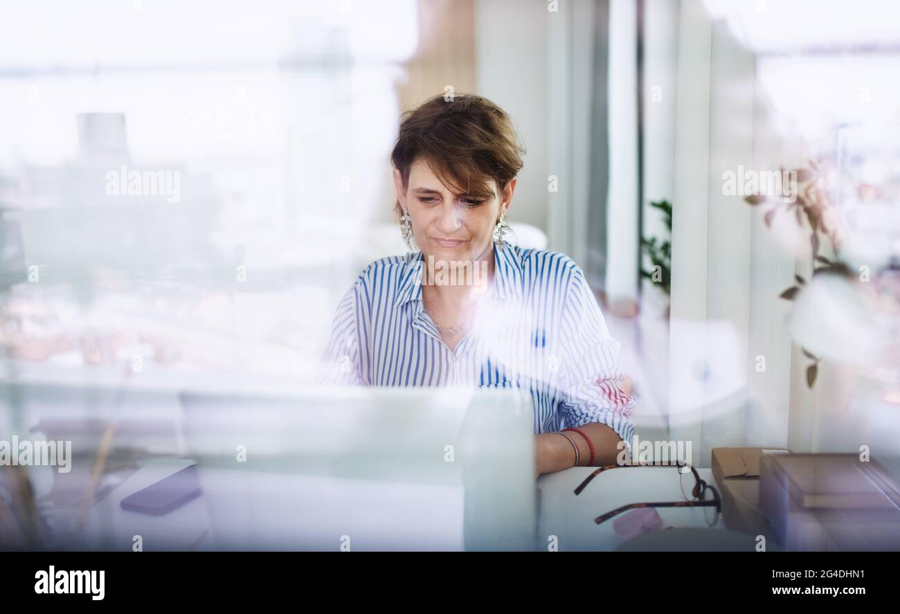 Mature woman with laptop working indoors in home office, small business concept. Shot through glass. Stock Photo