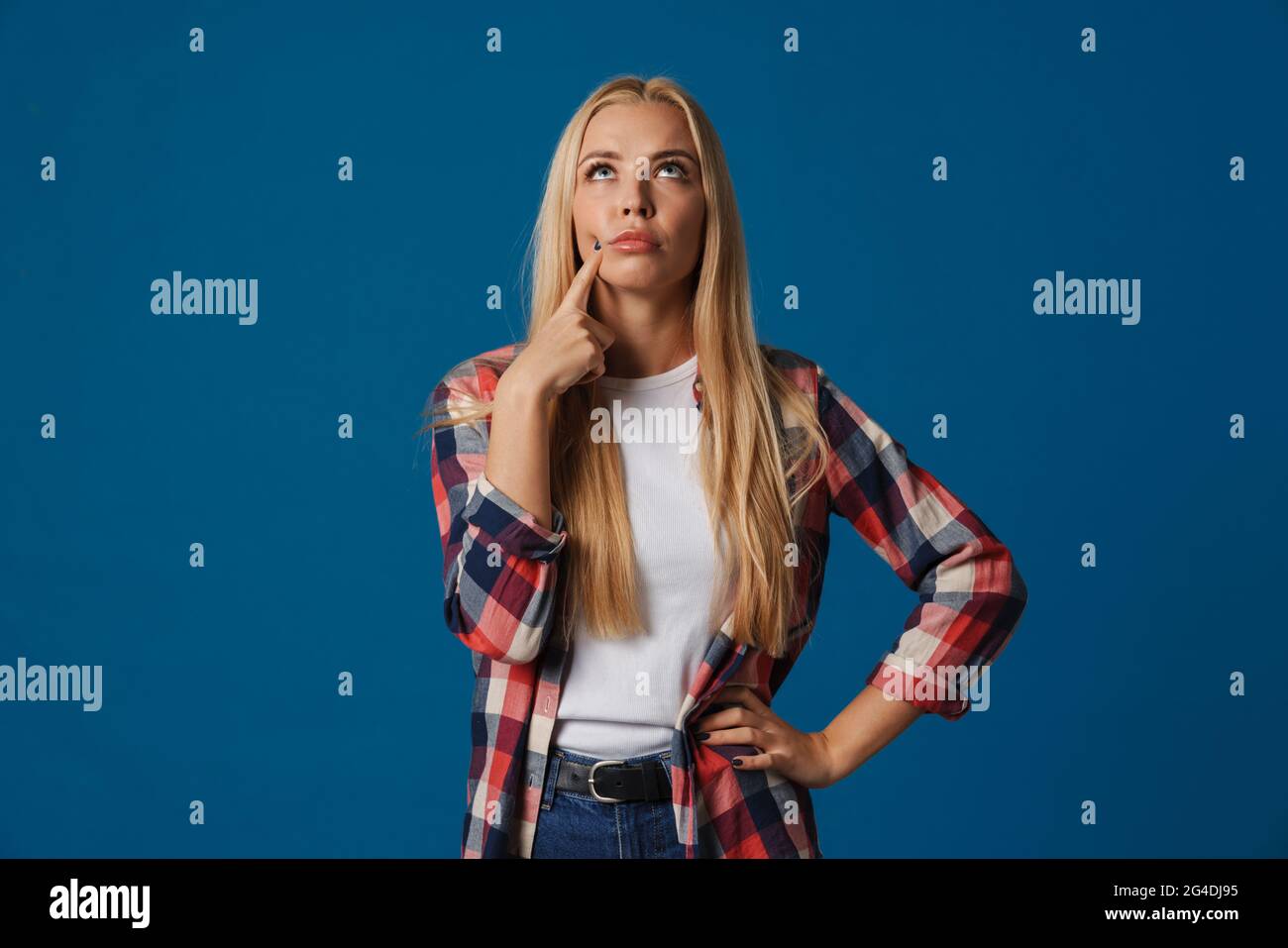 Brooding blonde girl pointing finger at her cheek and looking upward ...