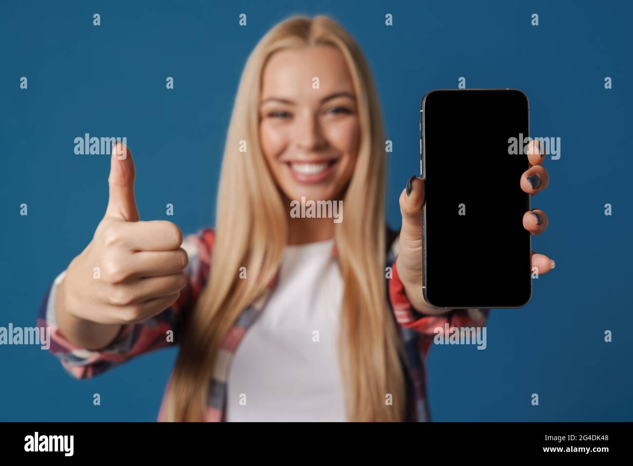 Blonde happy woman showing thumb up while showing mobile phone isolated over blue background Stock Photo