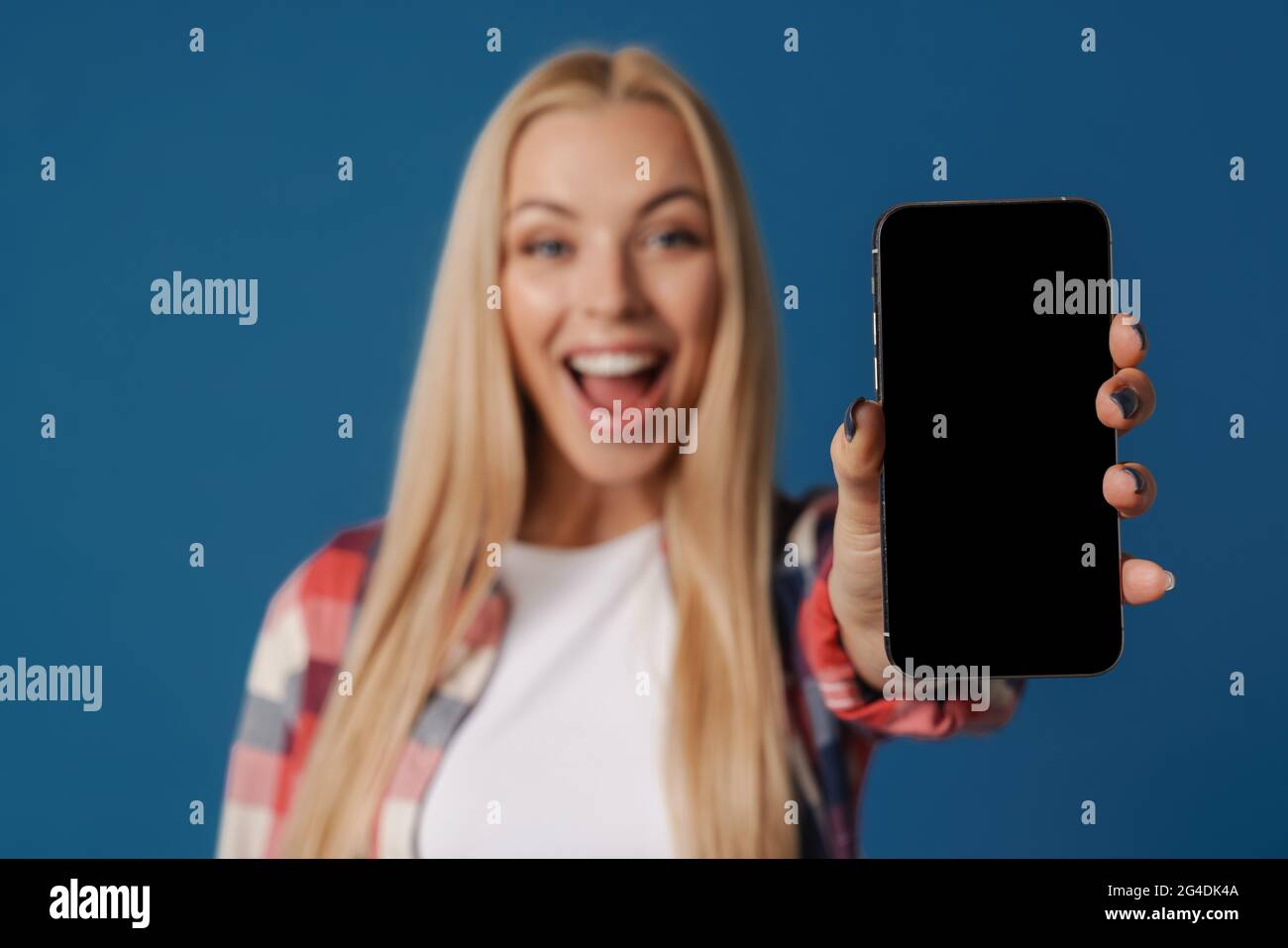 Blonde excited white woman smiling while showing mobile phone isolated over blue background Stock Photo