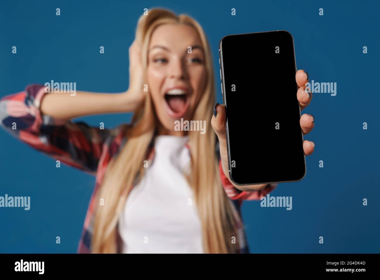 Blonde excited white woman exclaiming while showing mobile phone isolated over blue background Stock Photo