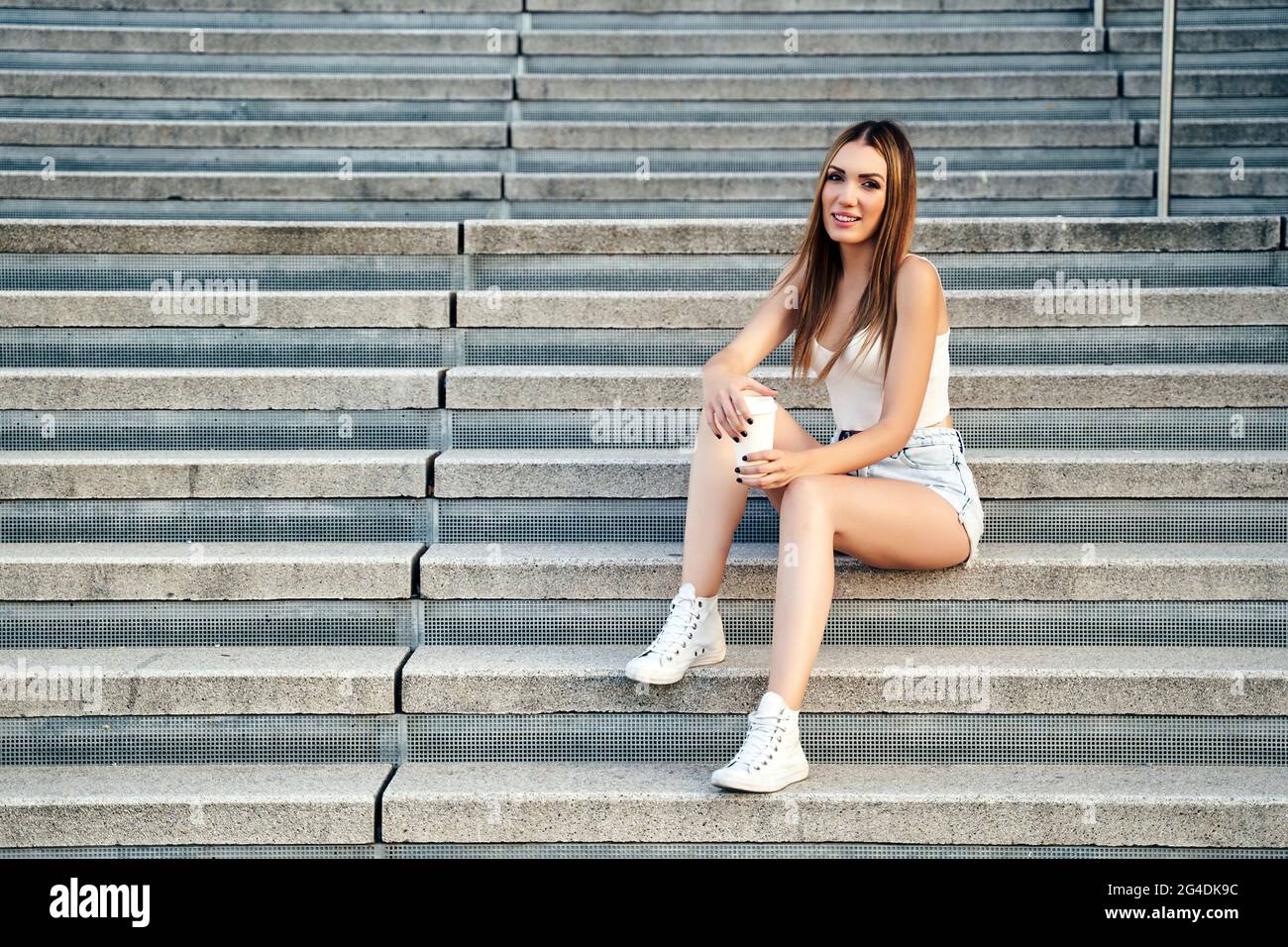 Beautiful girl, blonde with long hair, sitting on stairs, in summer clothes, drinking coffee. High quality photo Stock Photo