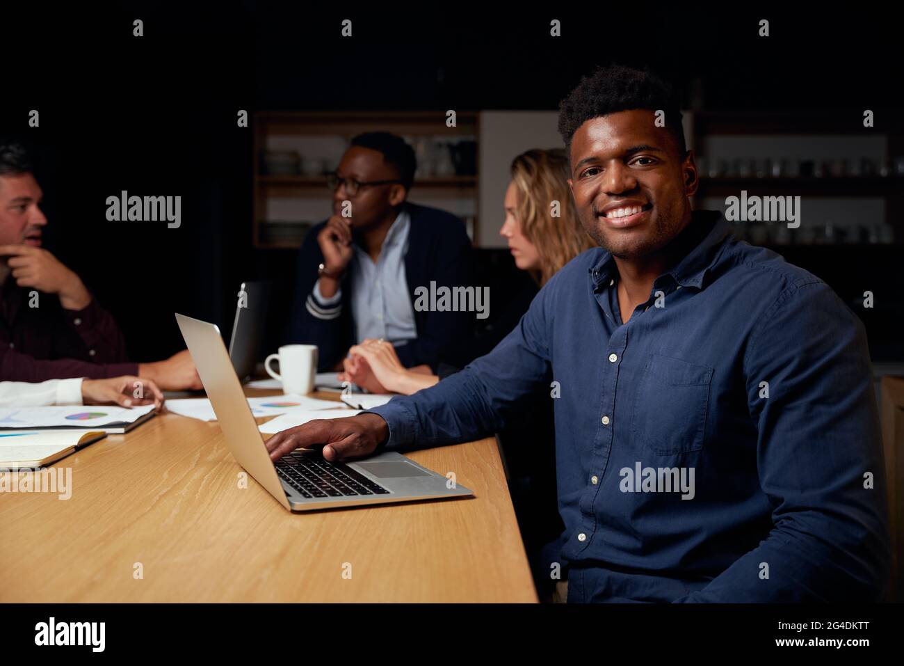 Portrait of smiling young african male team leader with laptop looking at camera while his colleague discussing project at background Stock Photo
