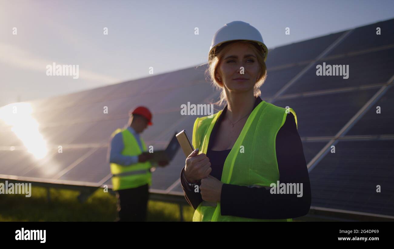 Portrait of a female solar engineer standing looking at the camera, another man near the panels and entering information into a laptop. Green energy Stock Photo