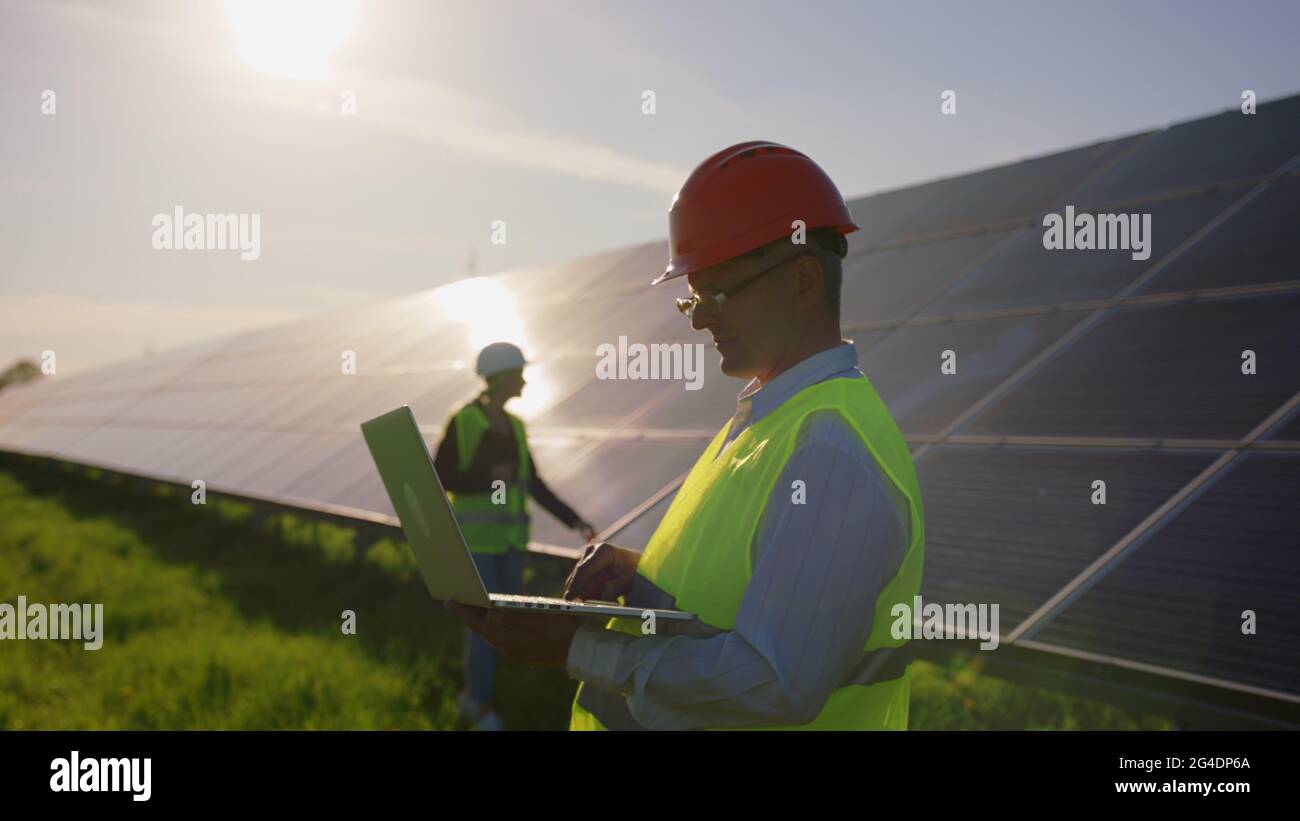 Portrait of a female solar engineer standing uses a tablet and looking at the camera, another man near the panels and entering information into a Stock Photo