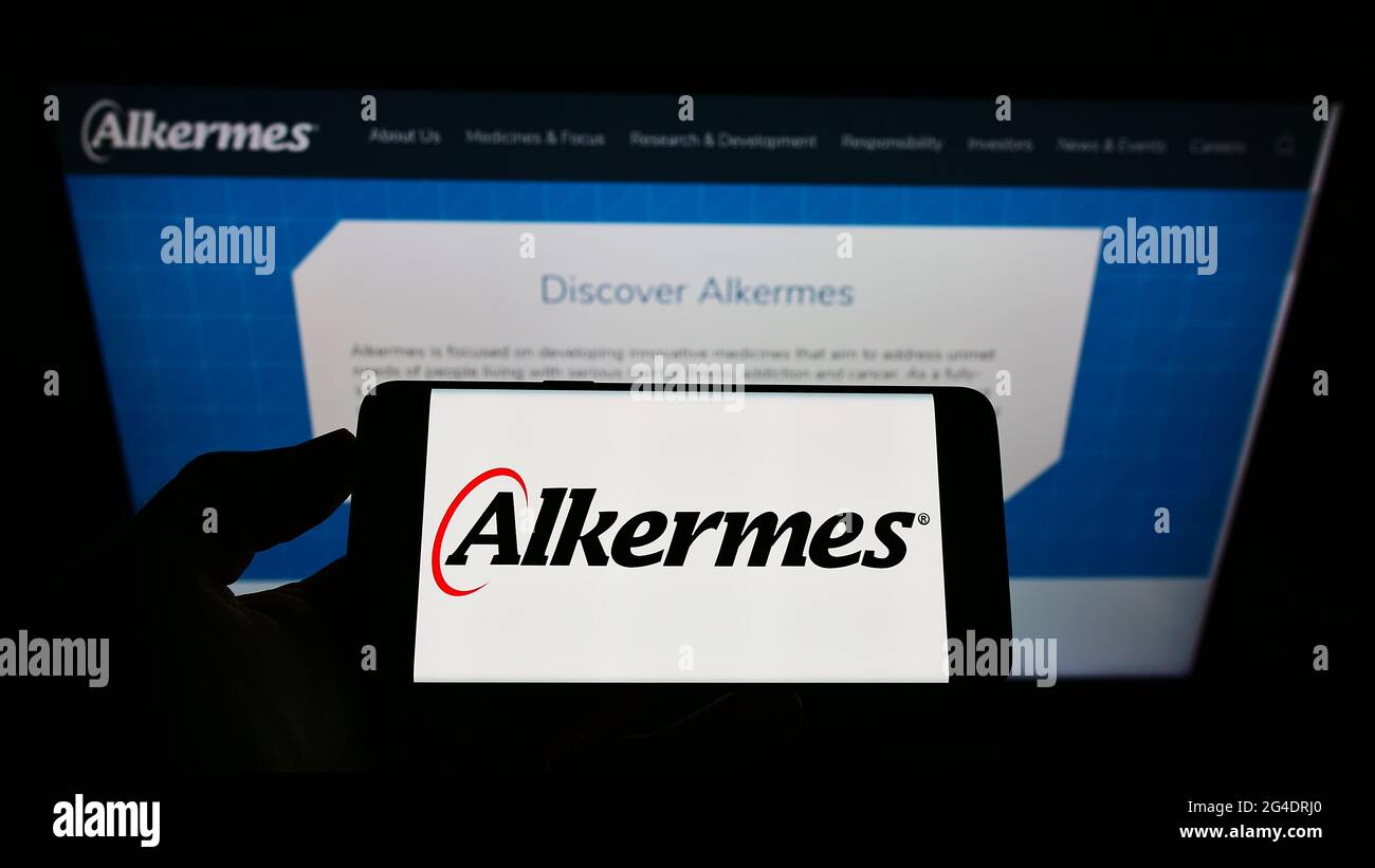 Person holding mobile phone with logo of Irish biopharmaceutical company Alkermes plc on screen in front of web page. Focus on phone display. Stock Photo